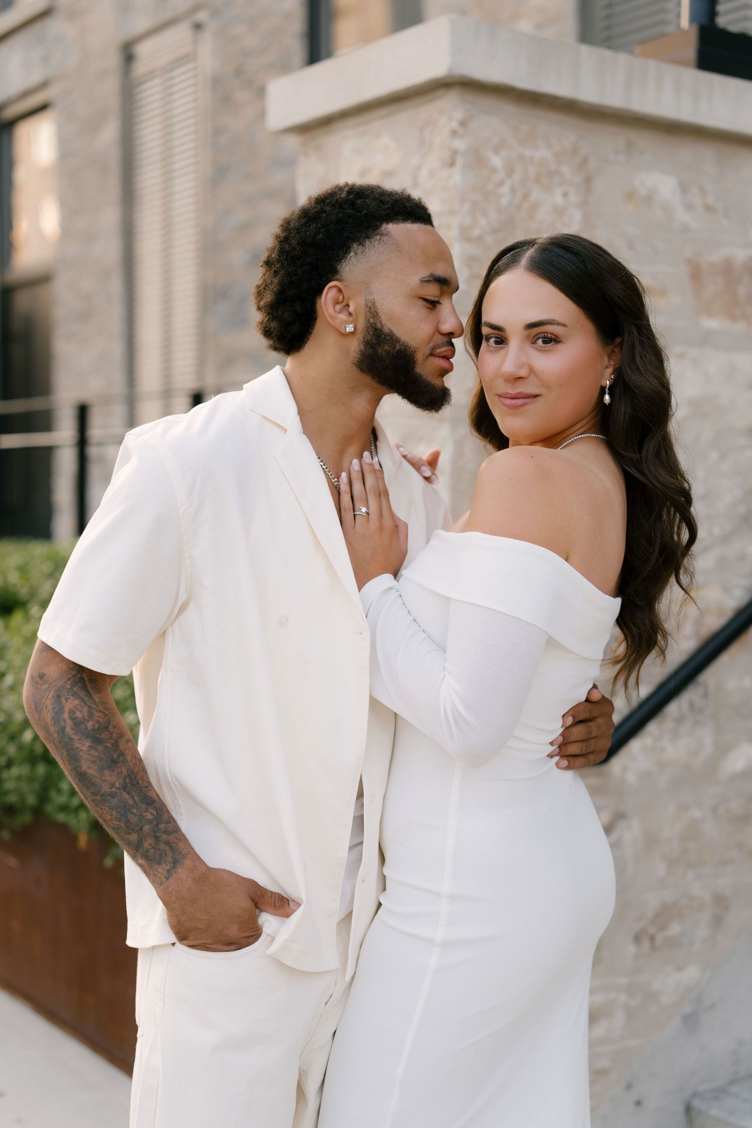 Couple in matching white outfits embrace outdoors, woman in off-shoulder gown touches man's chest.
