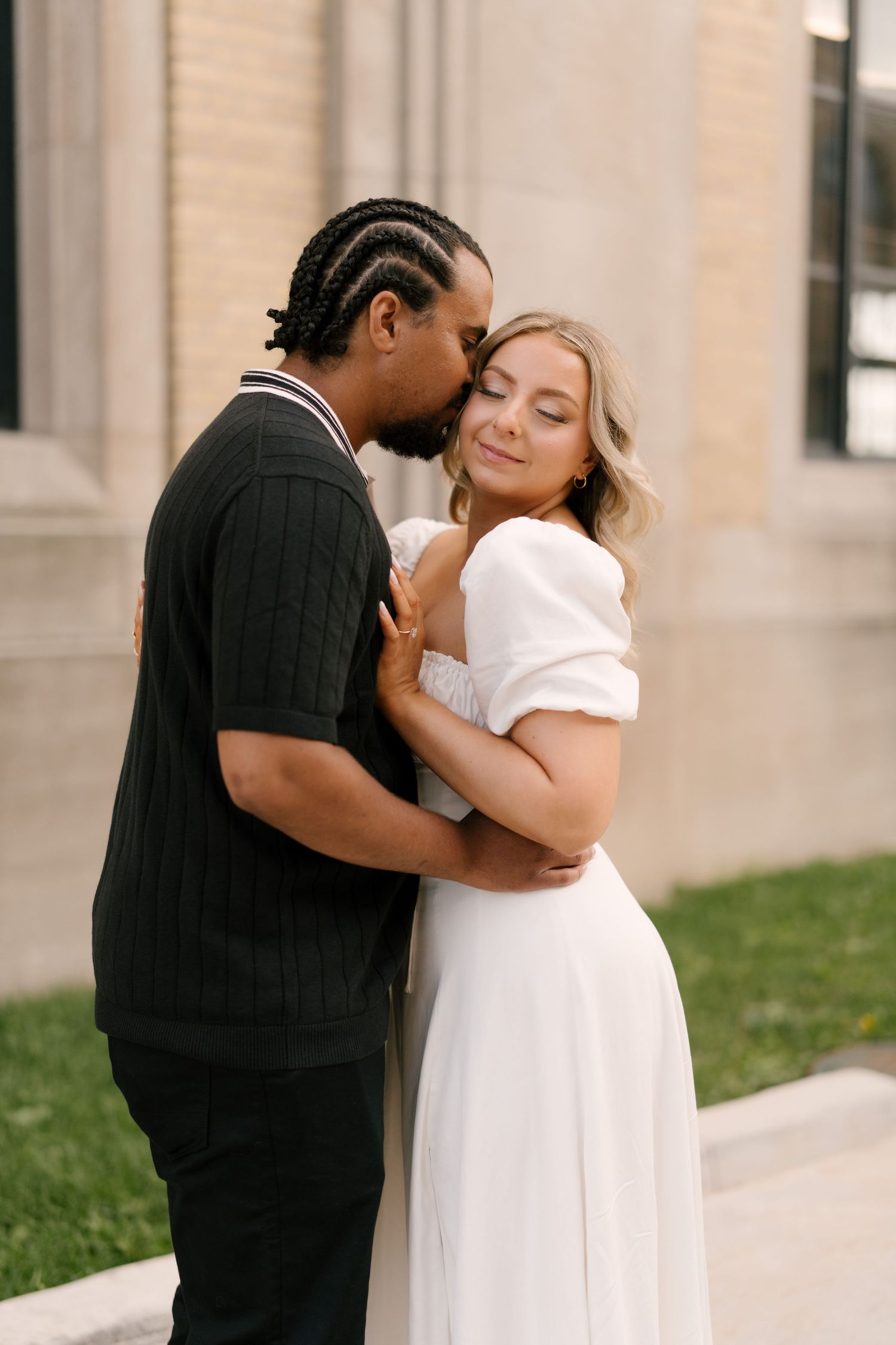 Interracial couple sharing a tender moment outdoors, man kissing woman in white dress near stone building.