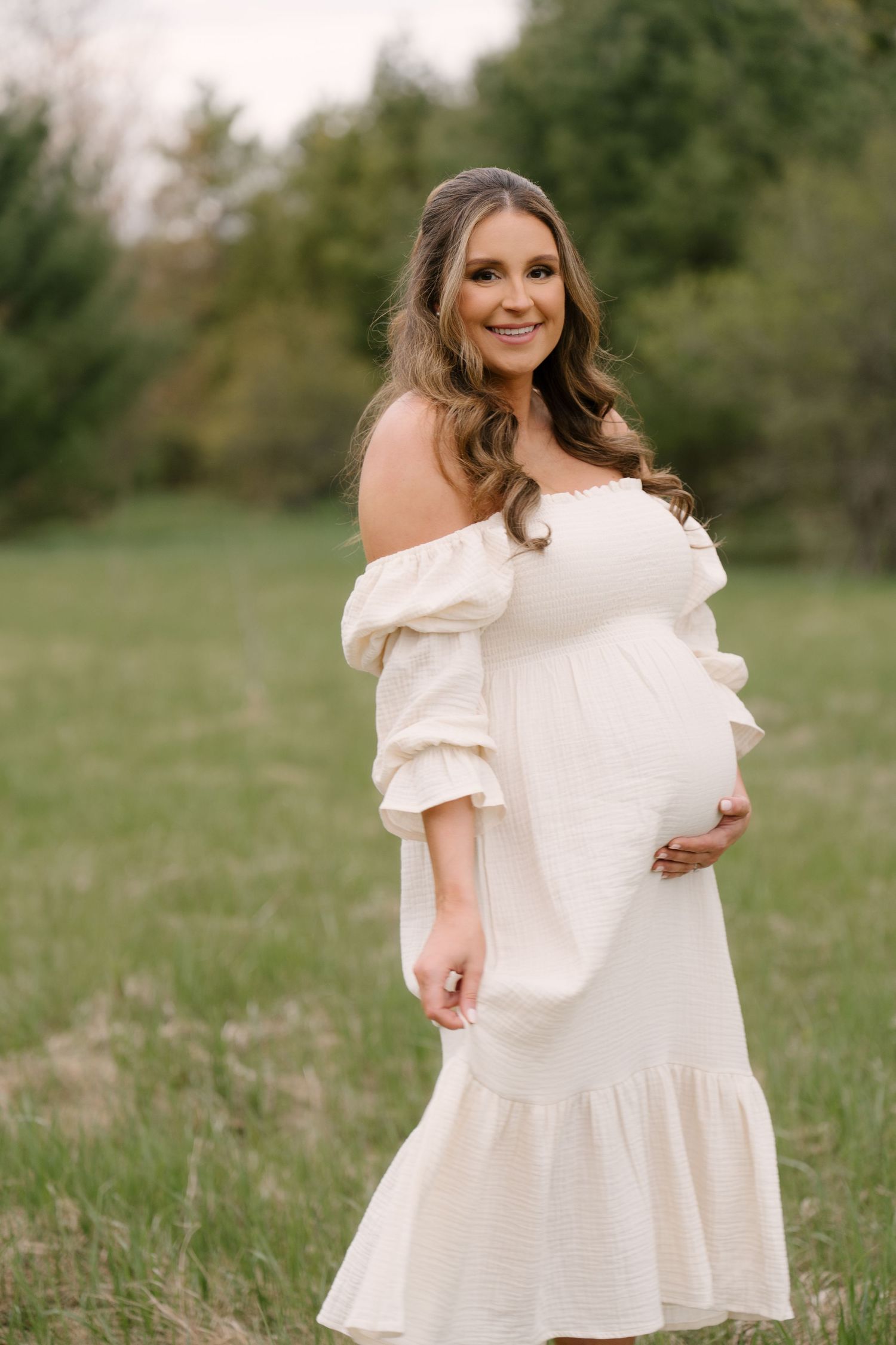 Elegant pregnant woman in white off-shoulder dress smiling in green field.