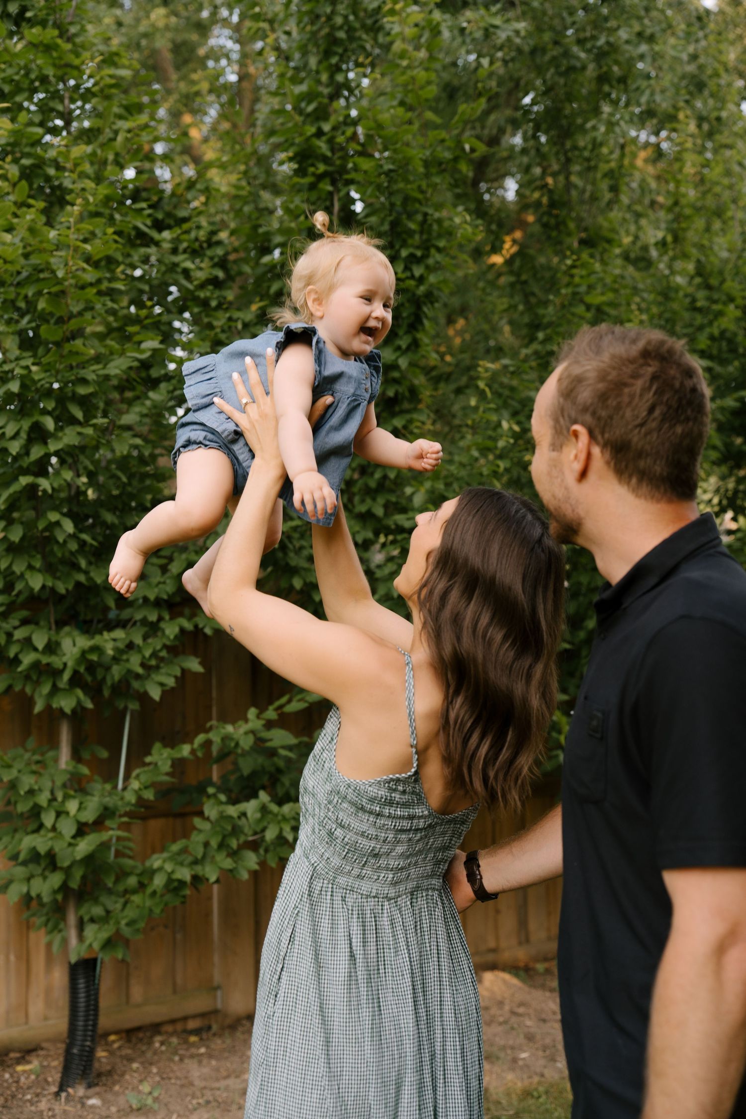 A mother lifts her laughing toddler in the air while father watches in a lush green backyard.