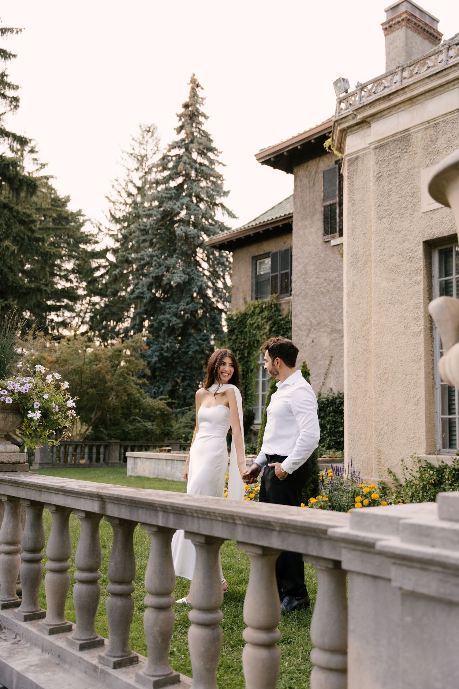 Elegant couple in wedding attire standing and laughing beside ornate stone balustrade at a grand estate garden.