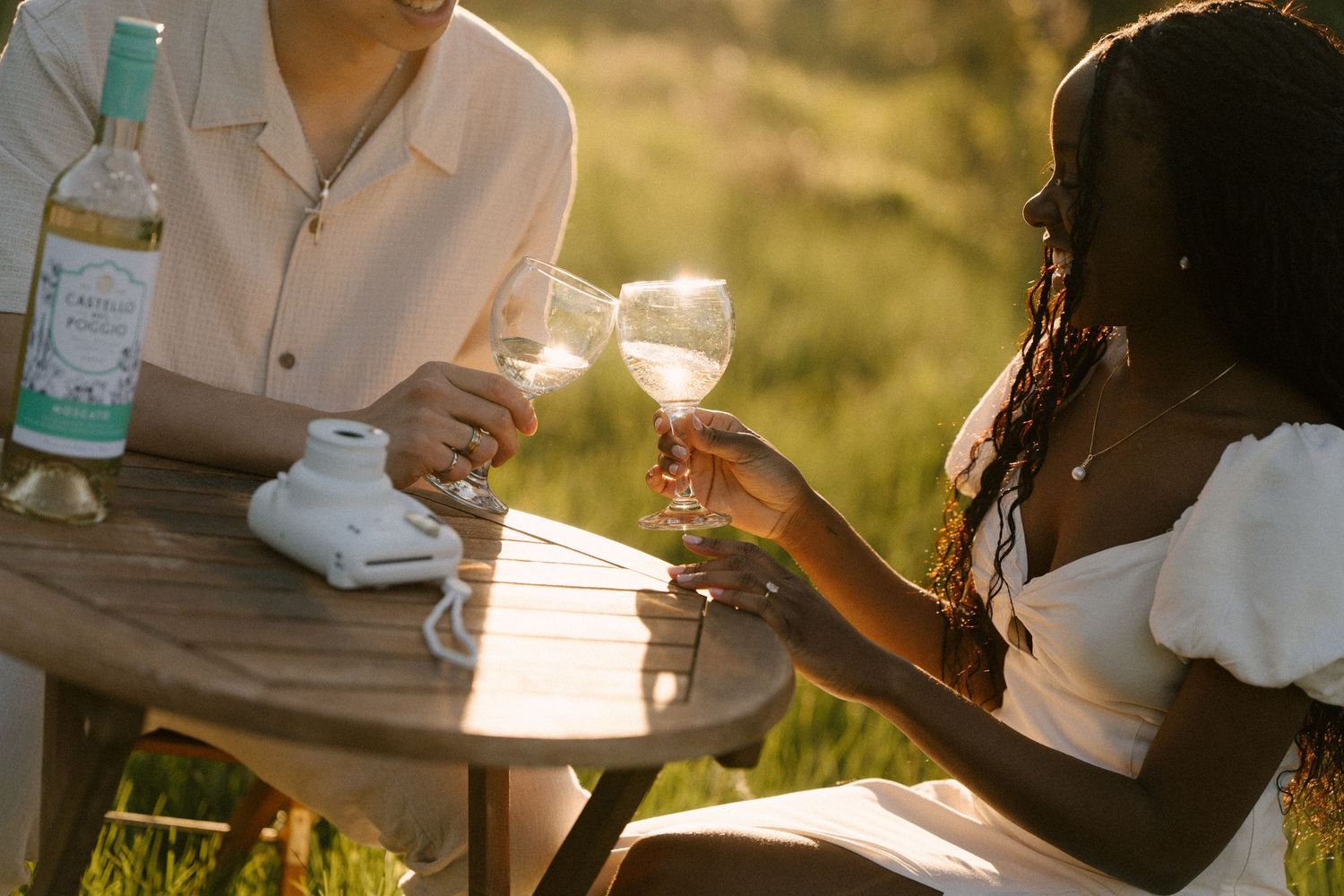 Couple clinking white wine glasses at outdoor table in golden sunset light.