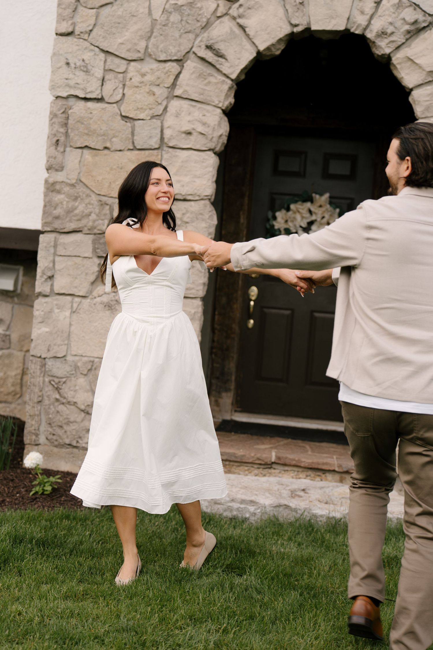 Smiling bride in white midi dress dancing with groom outside stone arch doorway.