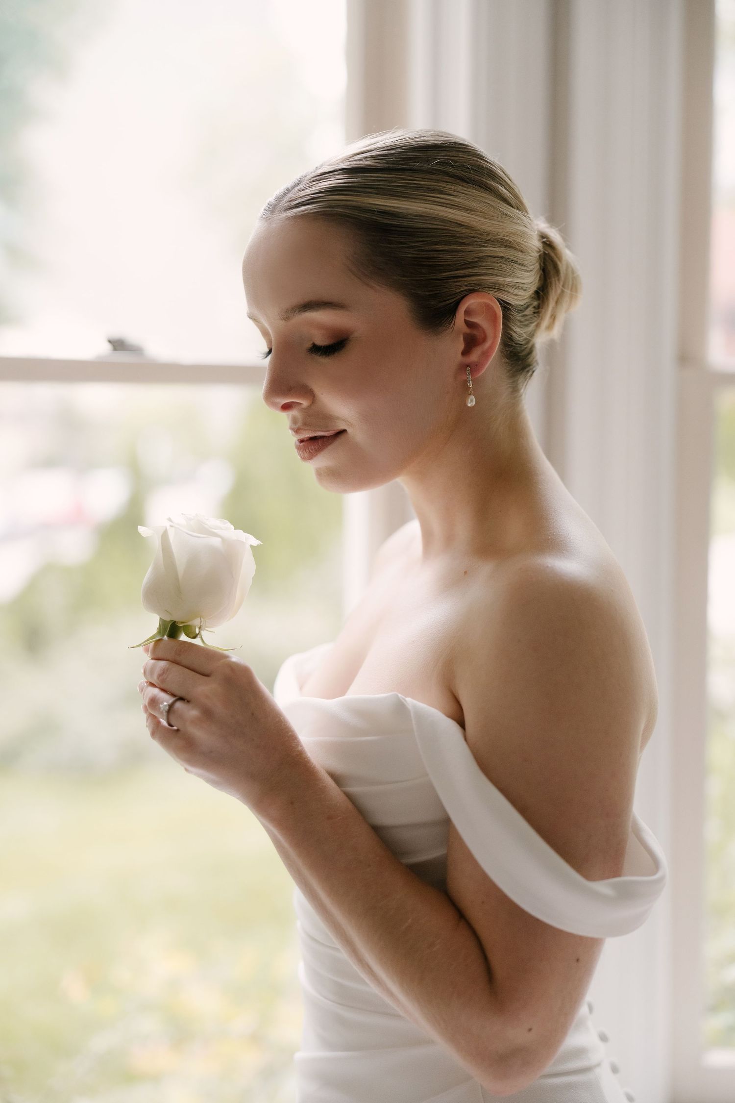 Bride in off-shoulder white gown holding a single white rose by a sunlit window.