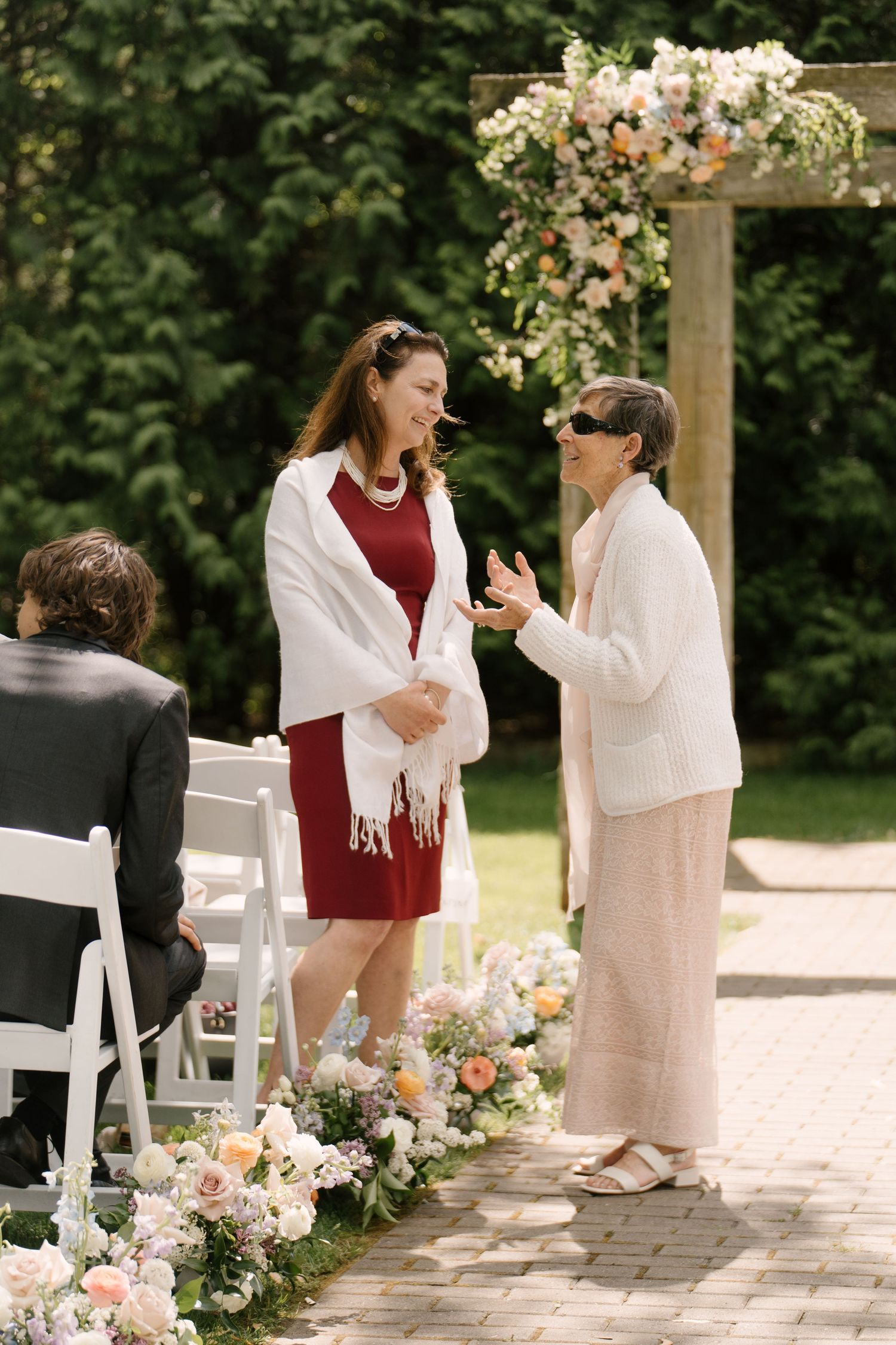 Two women in formal attire converse near floral arch and flower-lined aisle at outdoor ceremony at Roseville Estates wedding venue.