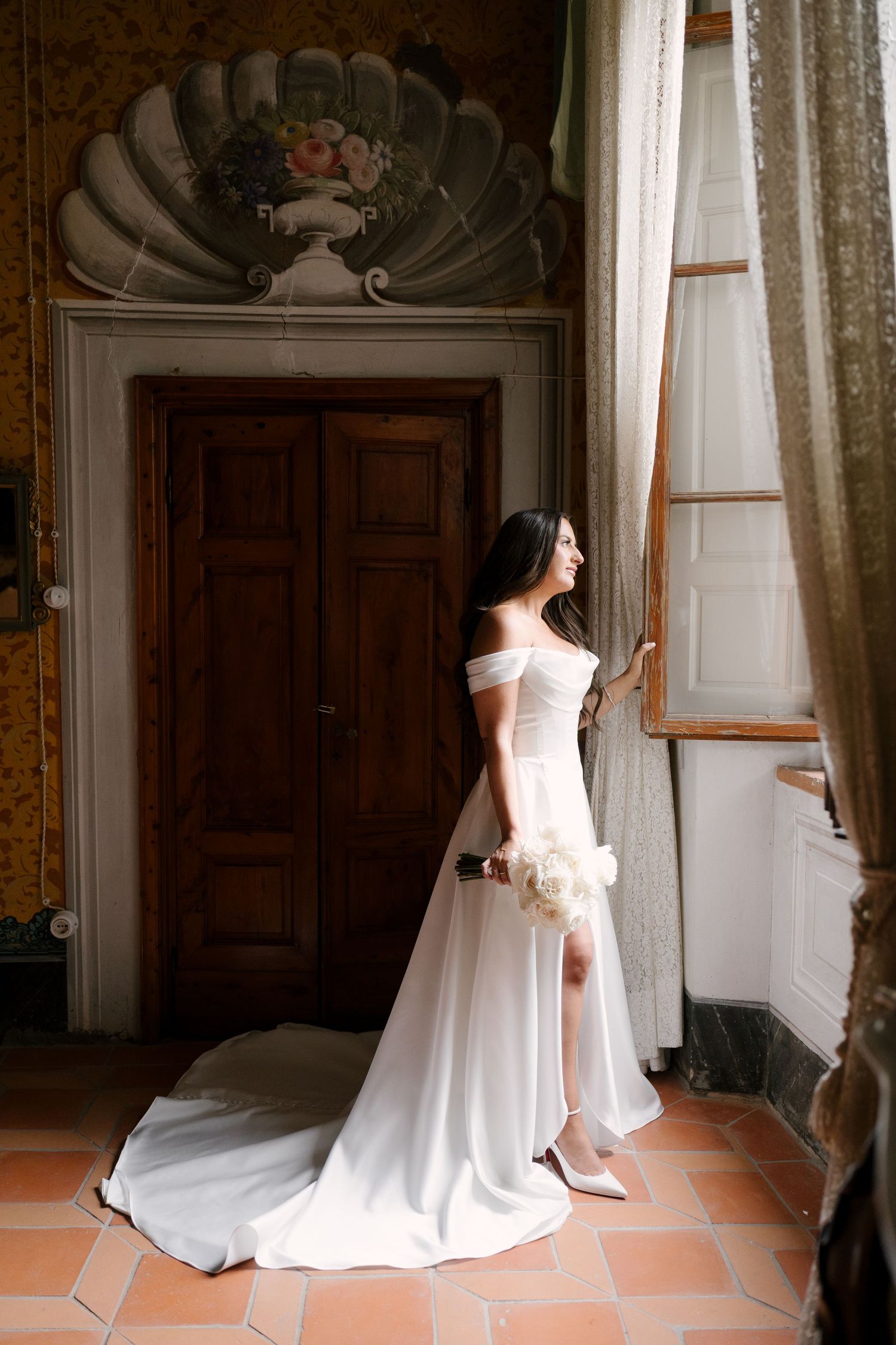 Elegant bride in white off-shoulder gown with train stands by ornate window in vintage villa interior.