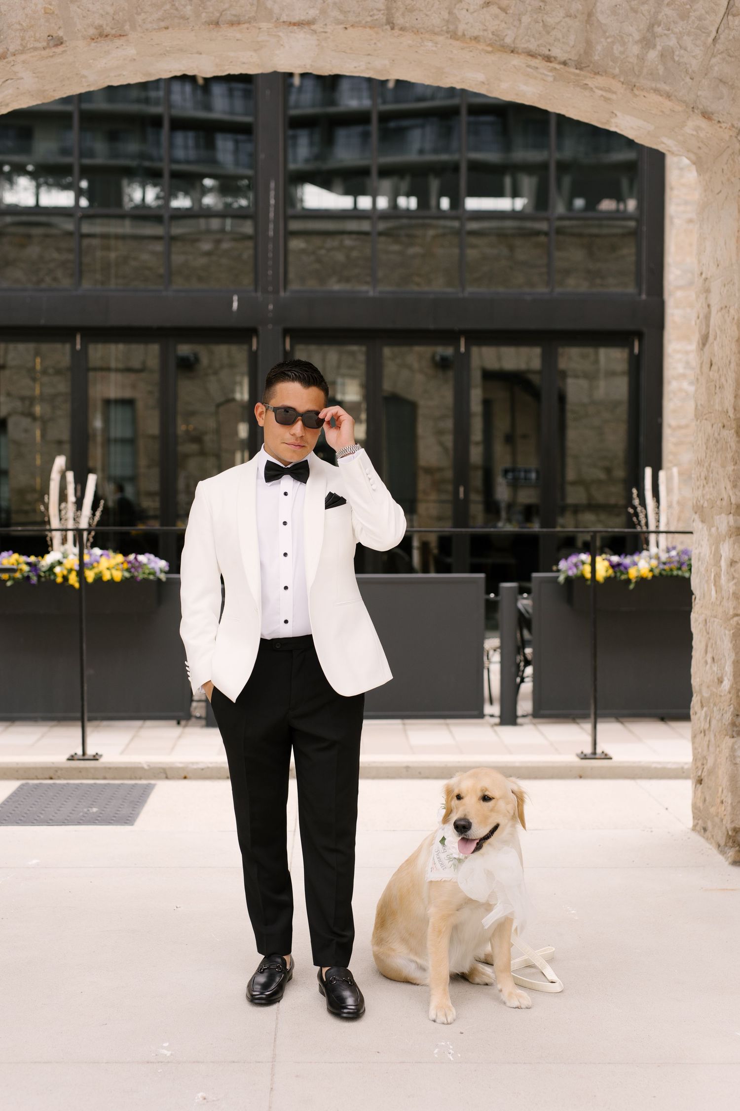 Groom in white tuxedo jacket adjusting sunglasses beside a golden retriever wearing a bow tie outside a stone building.