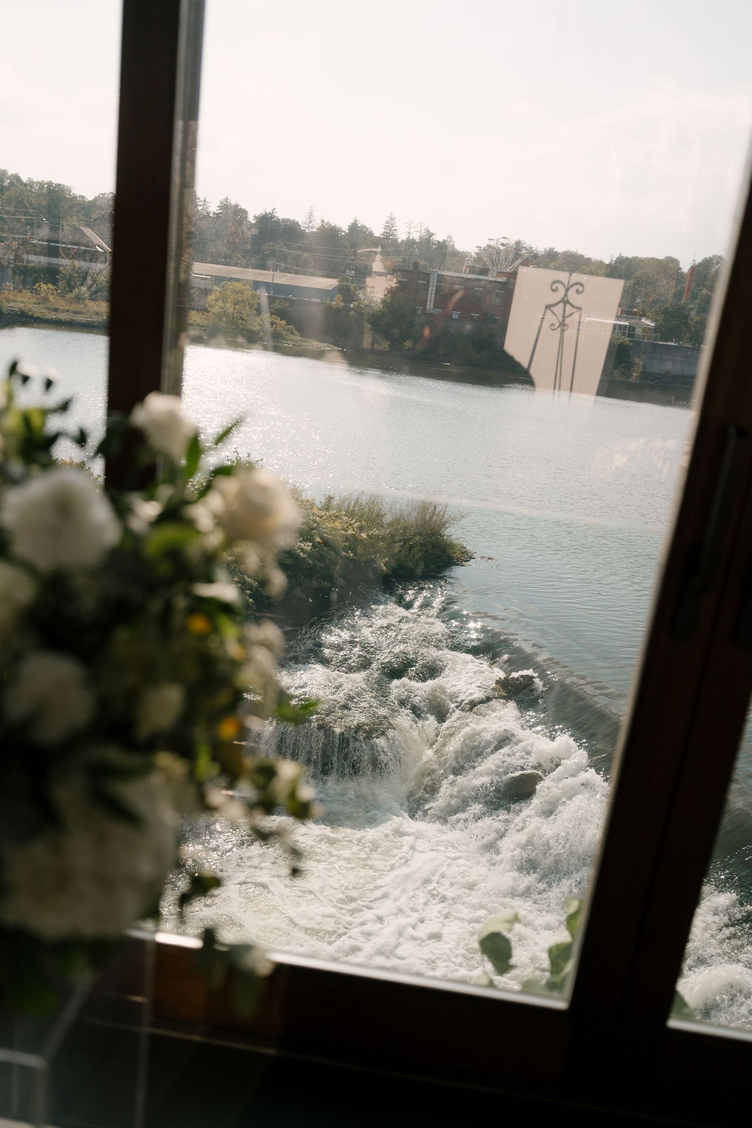 White wedding floral arrangement sits beside a window overlooking a rushing waterfall and river at Cambridge Mill.