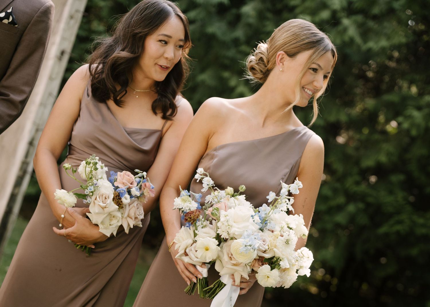 Two bridesmaids in taupe one-shoulder gowns holding white floral bouquets at an outdoor wedding ceremony.