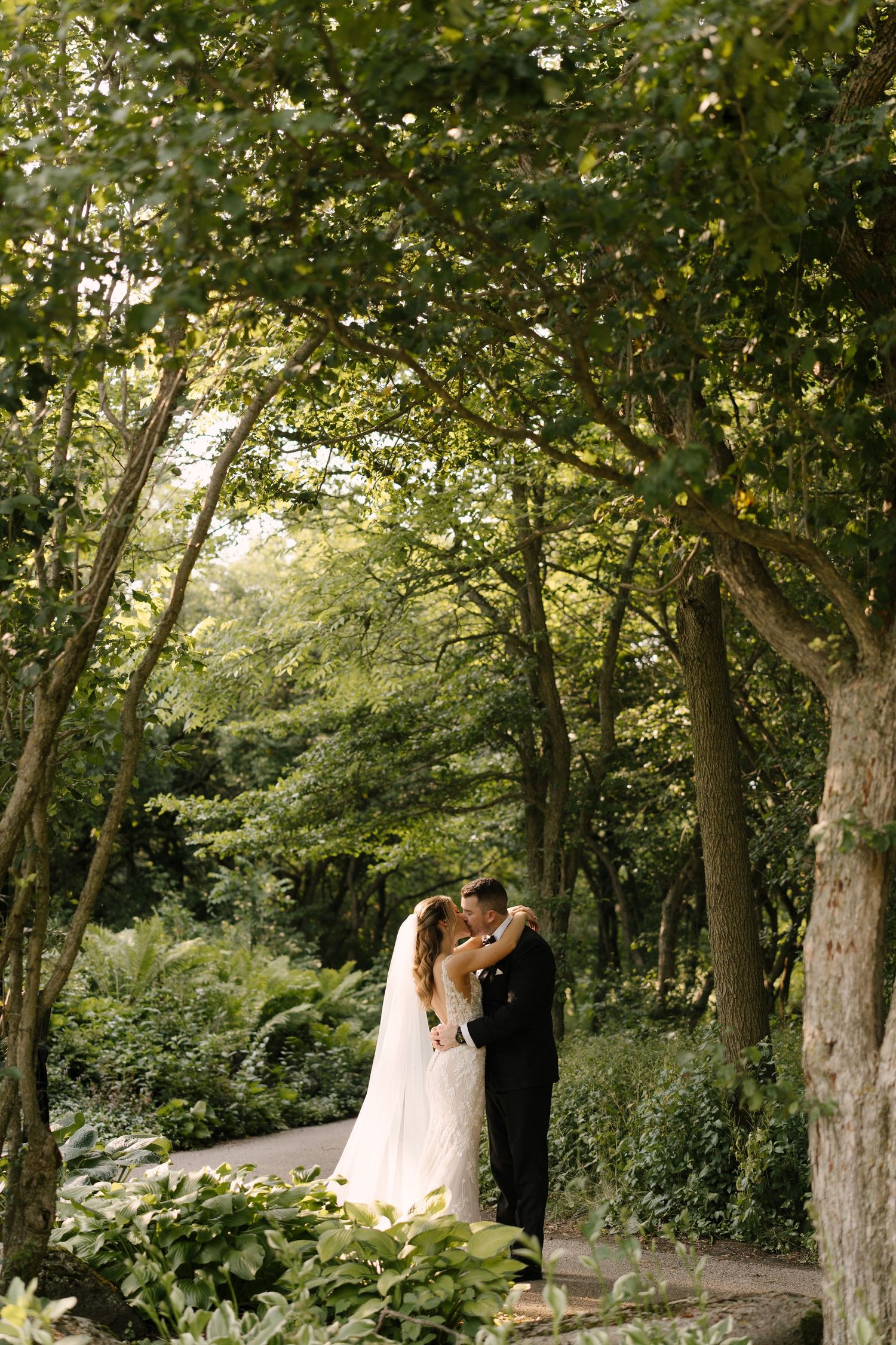 Bride and groom kissing on a tree-lined garden path with lush green foliage at St Marys venue.