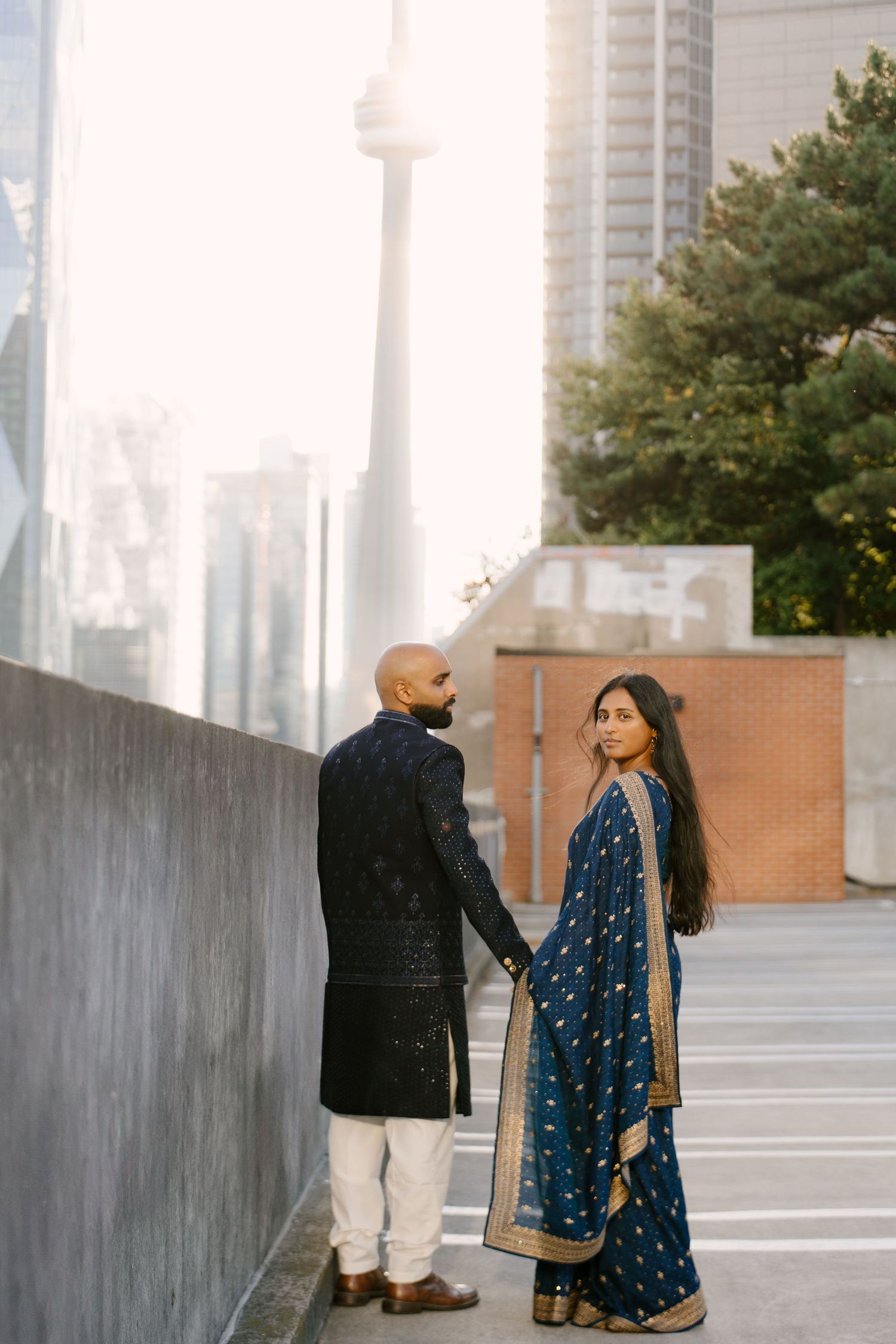 Indian couple in blue traditional attire hold hands with CN Tower visible behind them.