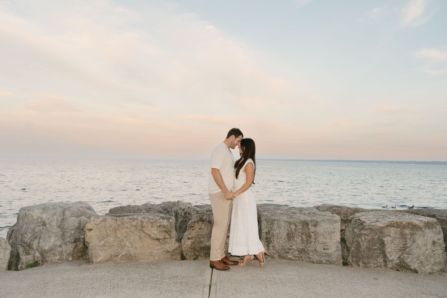 Couple holding hands facing each other on rocky beach wall at pastel sunset with calm ocean behind.