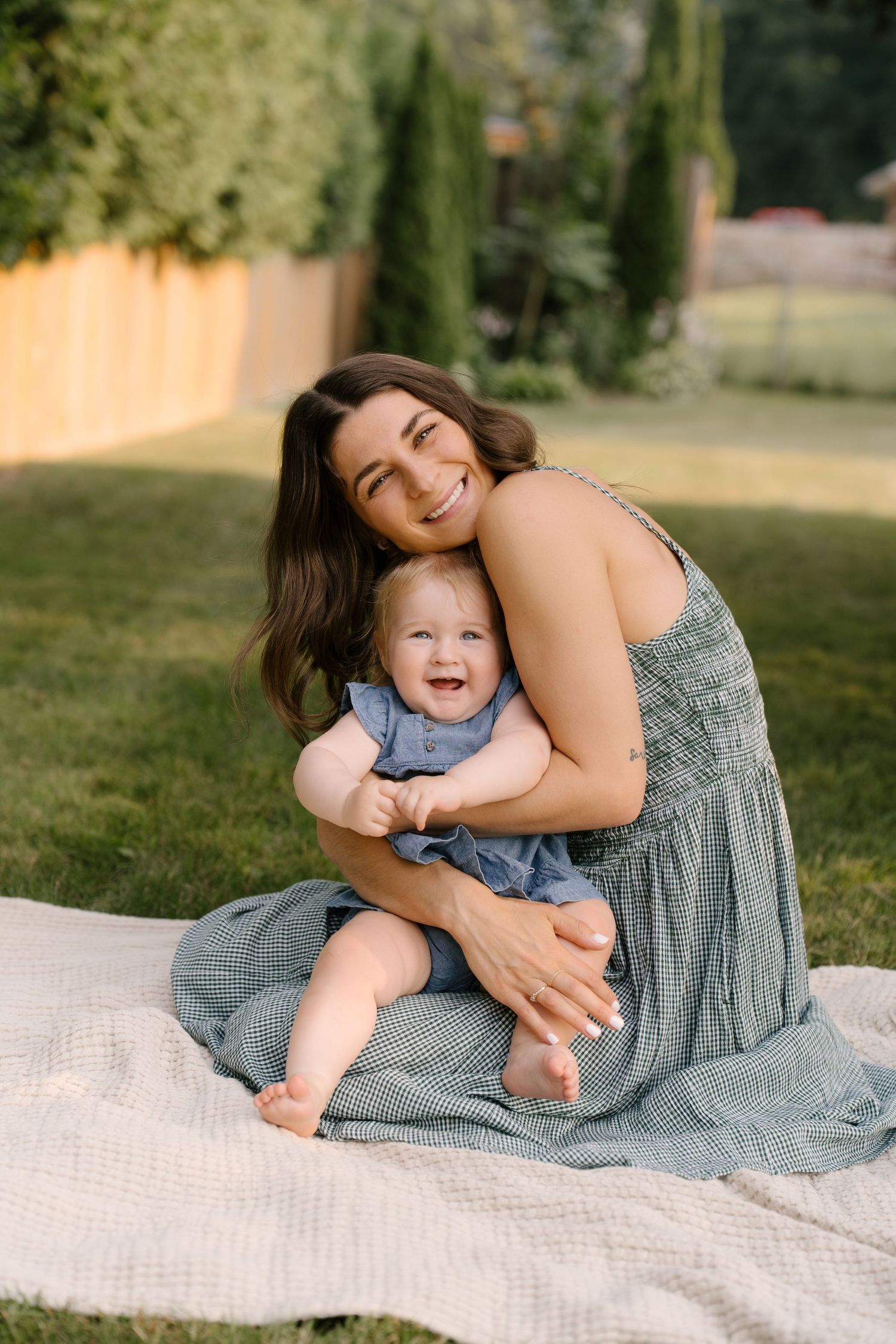 A smiling brunette mother hugs her laughing baby girl in a blue dress on a blanket in a sunny yard.