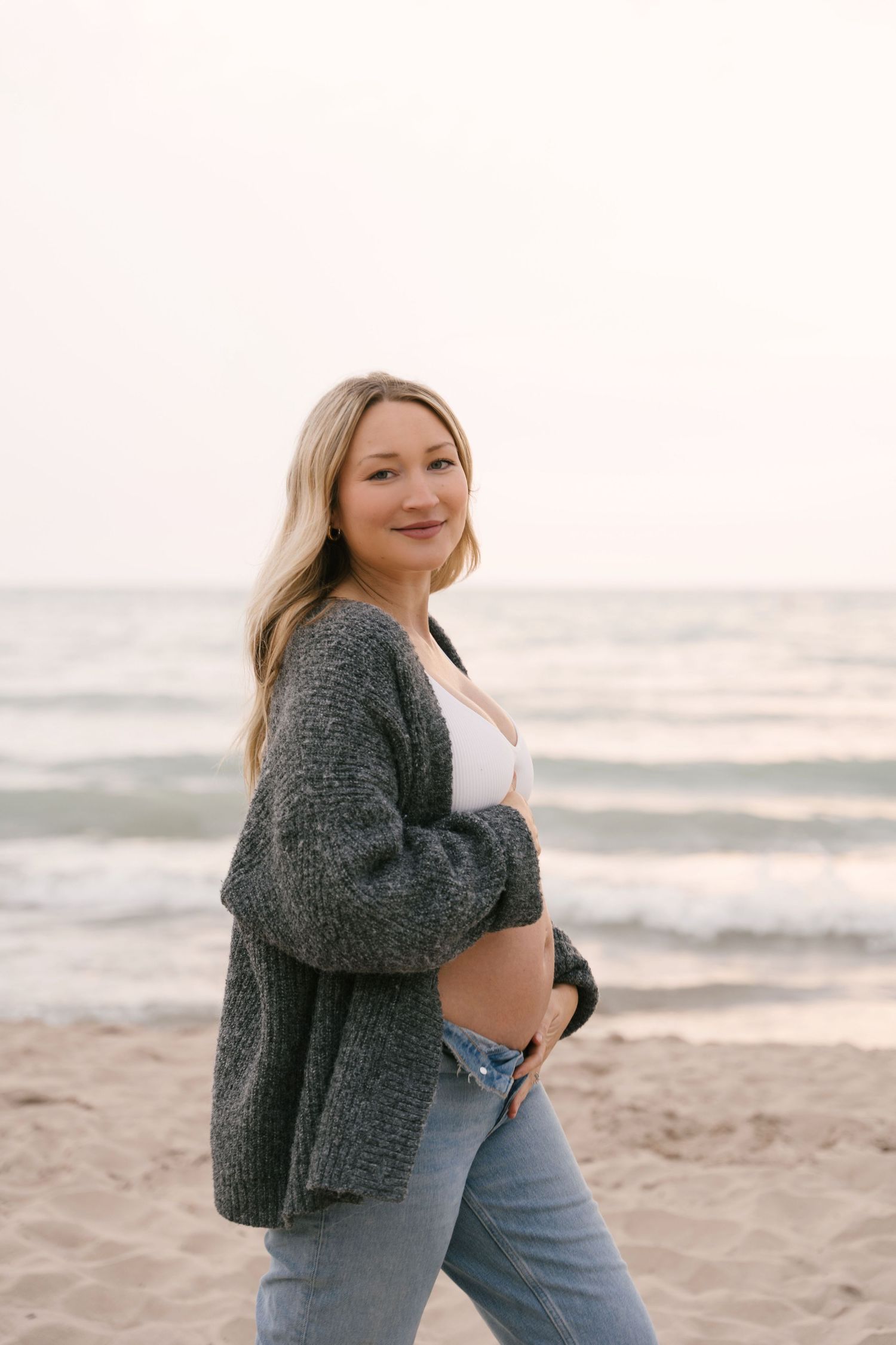 Confident pregnant woman in gray cardigan smiling at camera while walking on sandy beach.