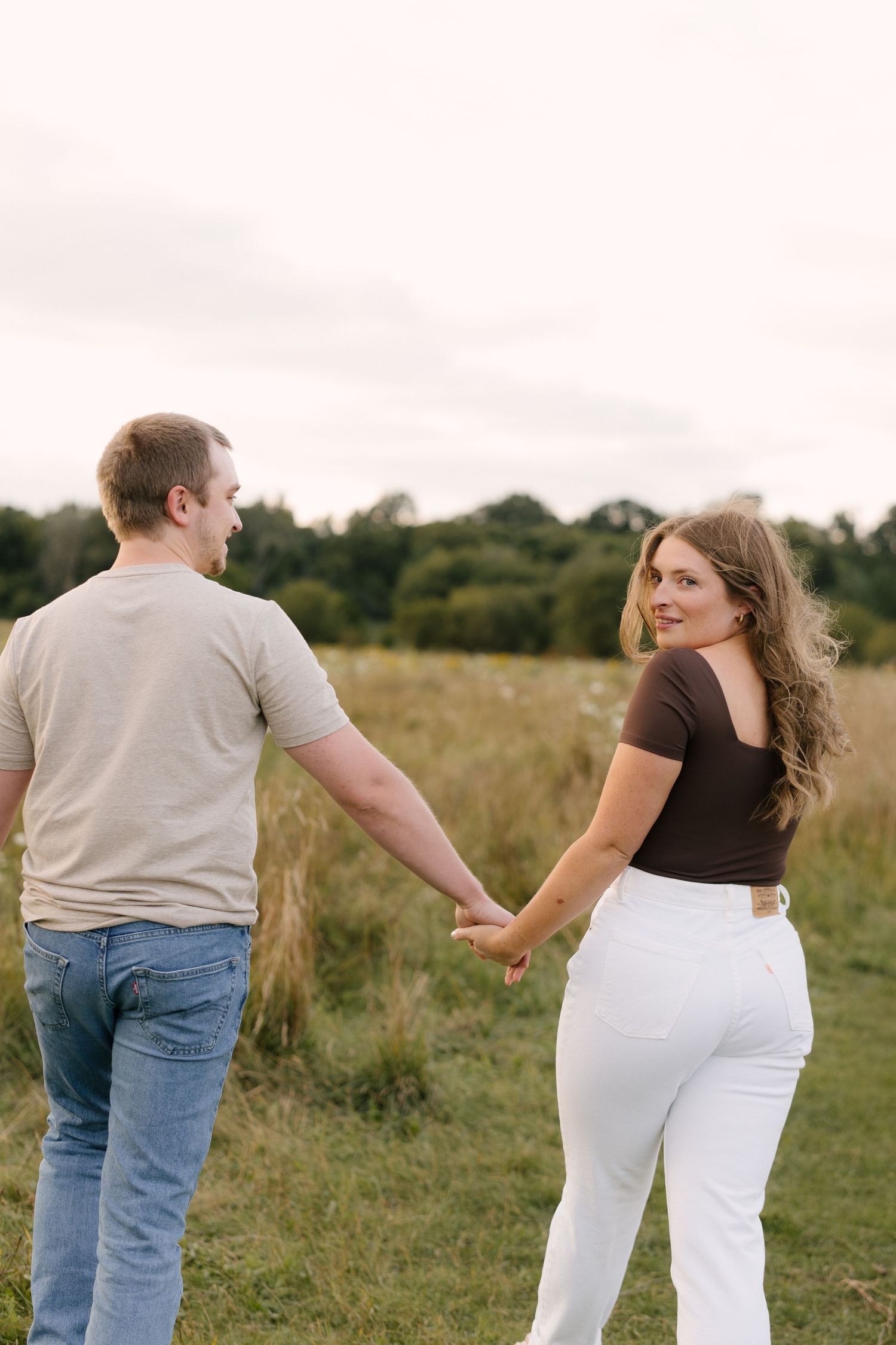 Woman smiling over her shoulder while holding hands with man walking through a meadow.