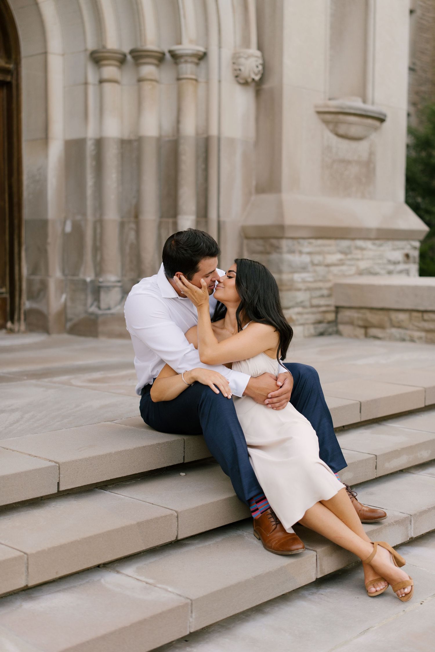 Couple sharing a kiss while sitting on stone church steps in romantic engagement photo.