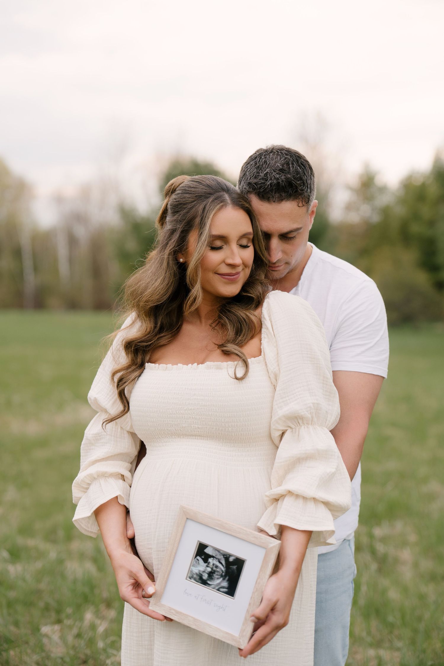 Couple tenderly holding framed ultrasound photo together in a green field.