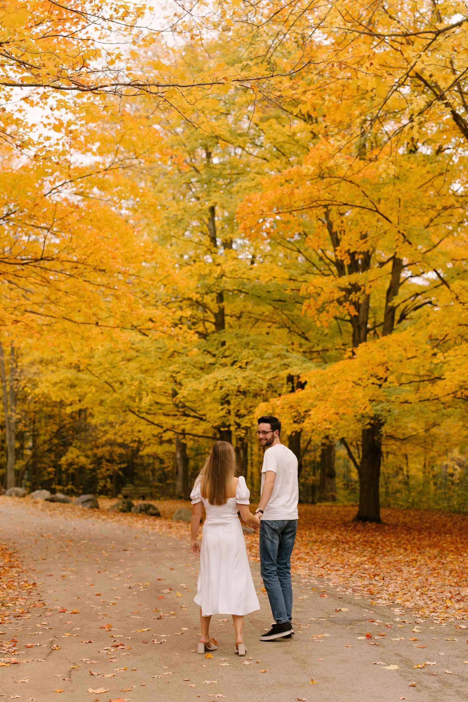 A couple holds hands while walking along a scenic path lined with golden autumn foliage.