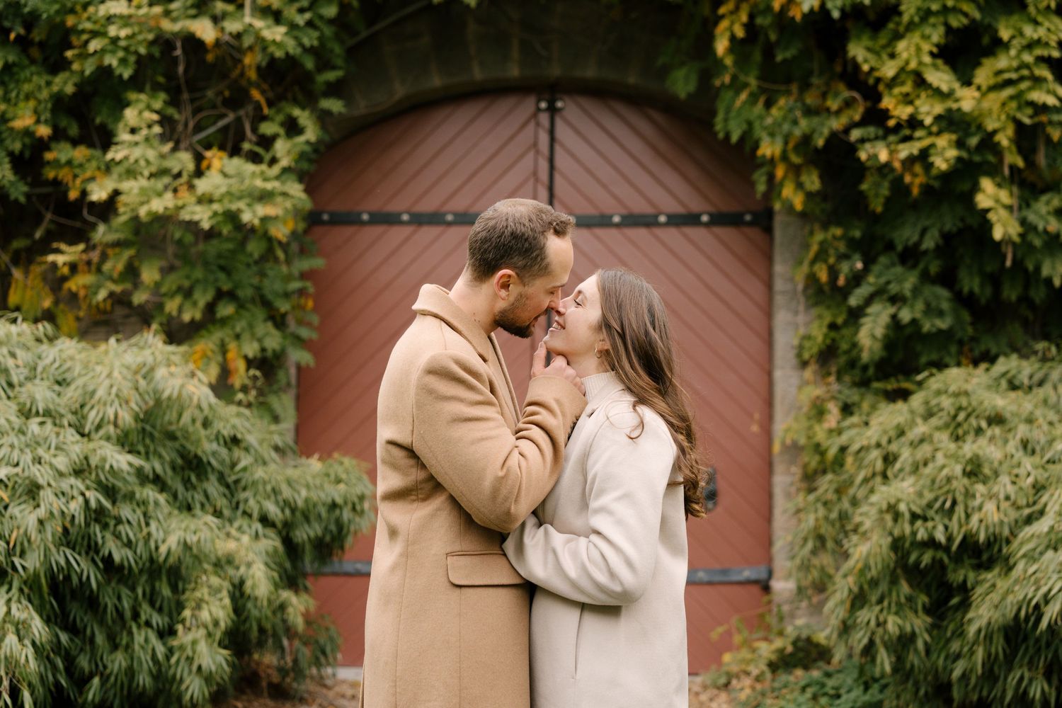 Couple sharing a tender moment, nearly kissing, wearing fall coats before a rustic red barn door with ivy.