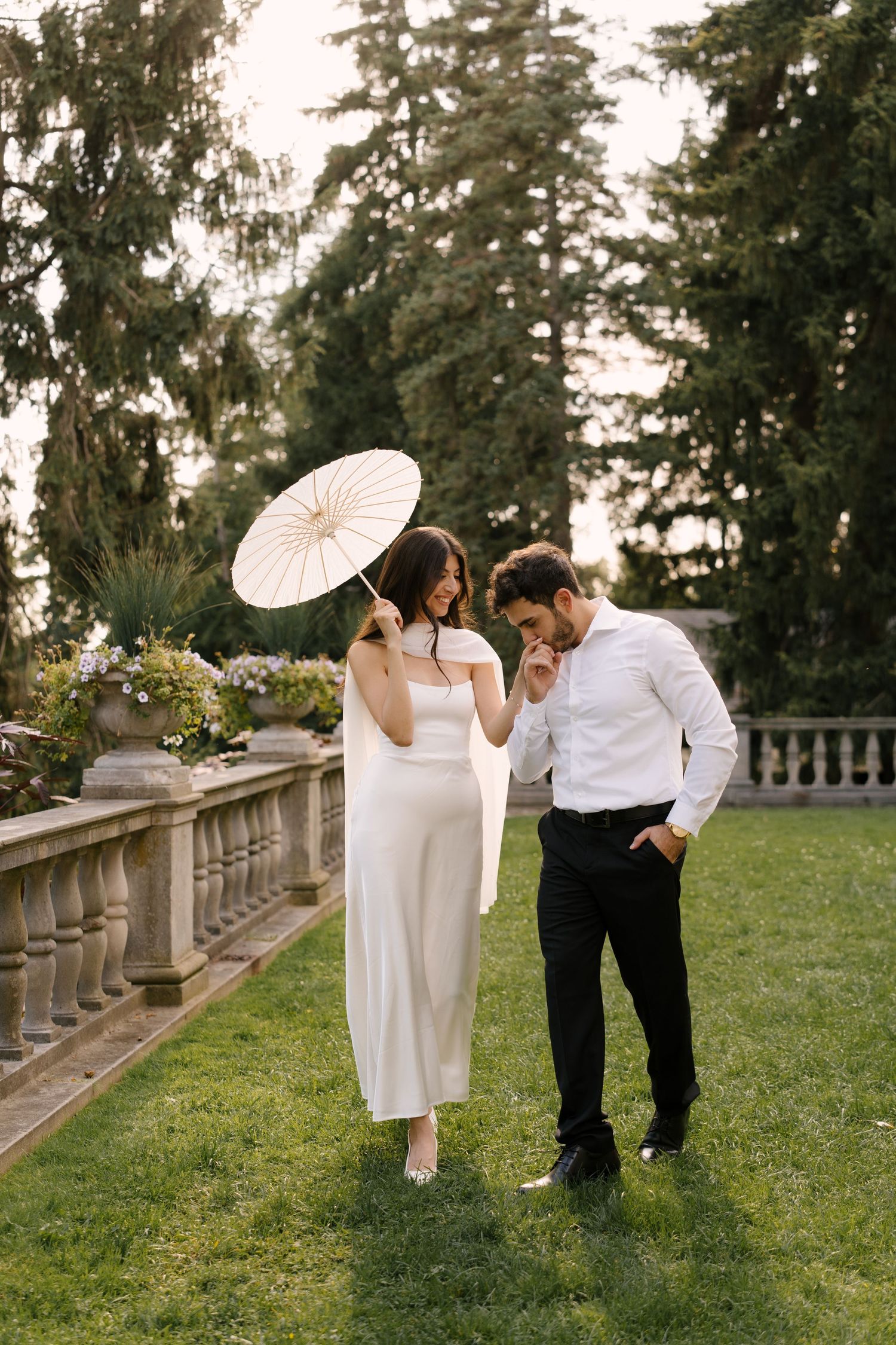 Smiling bride twirling white parasol beside laughing groom in white shirt on garden lawn at wedding venue.