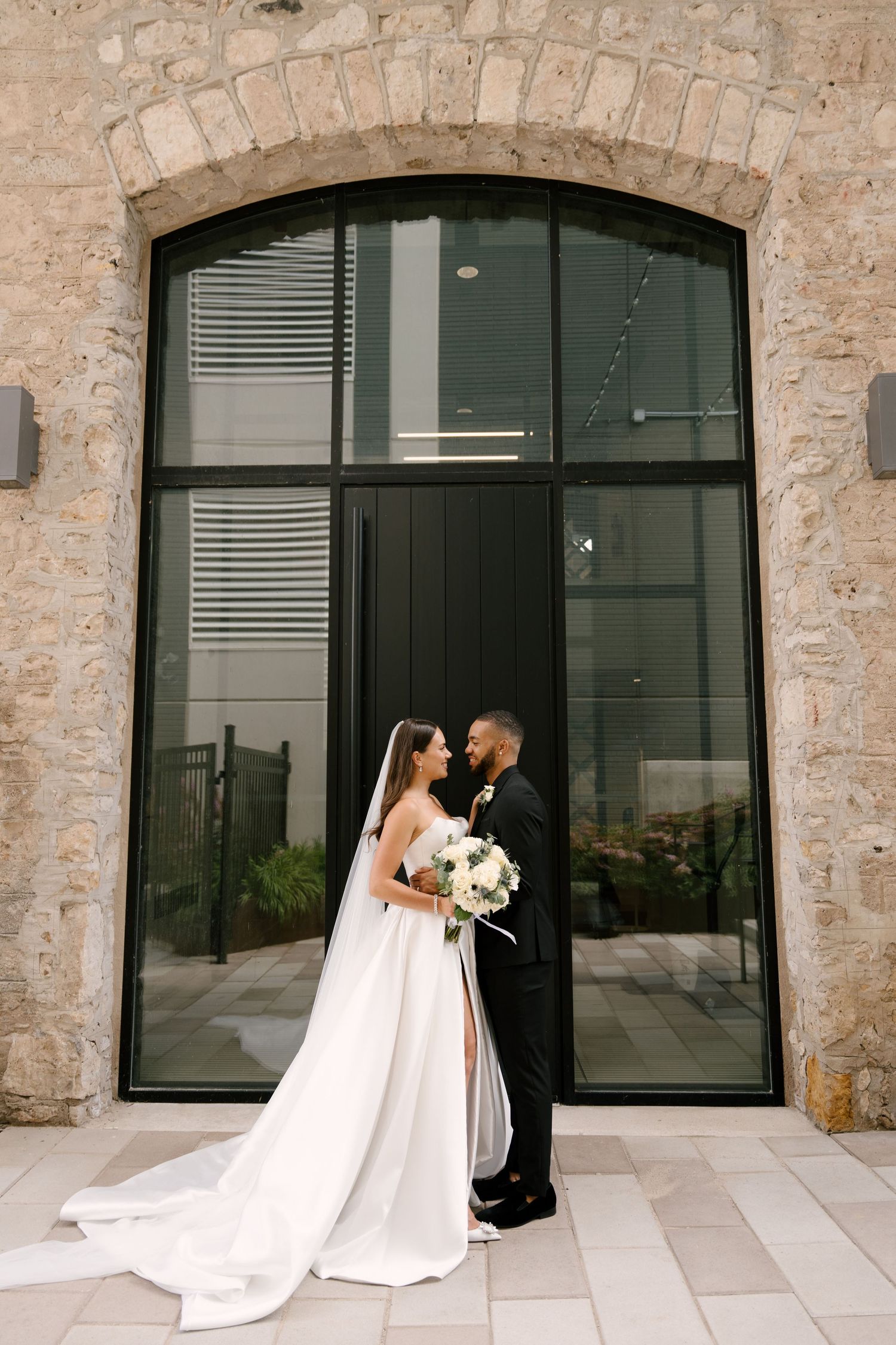 Bride and groom stand facing each other before a stone arch and large black door.