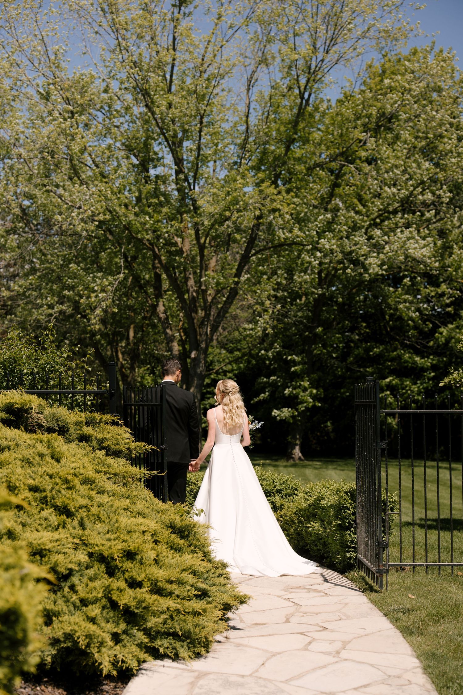 Bride and groom walk hand in hand along garden stone path surrounded by lush greenery at Roseville Estates.