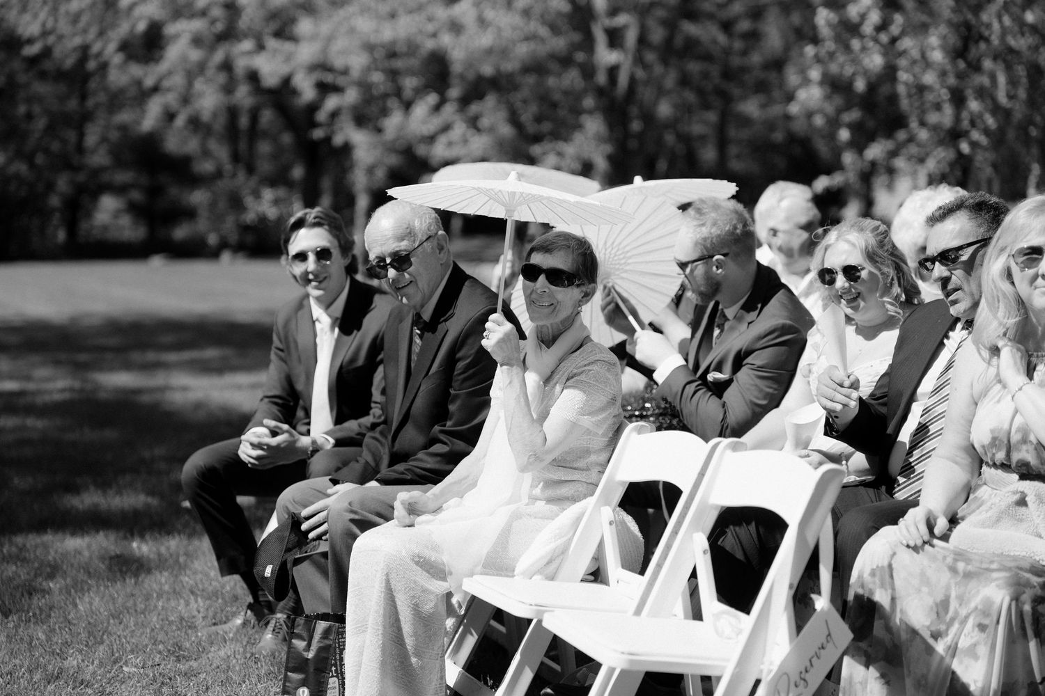 Wedding guests seated outdoors in sunlight, one shielding sun with a white parasol at Roseville Estates.