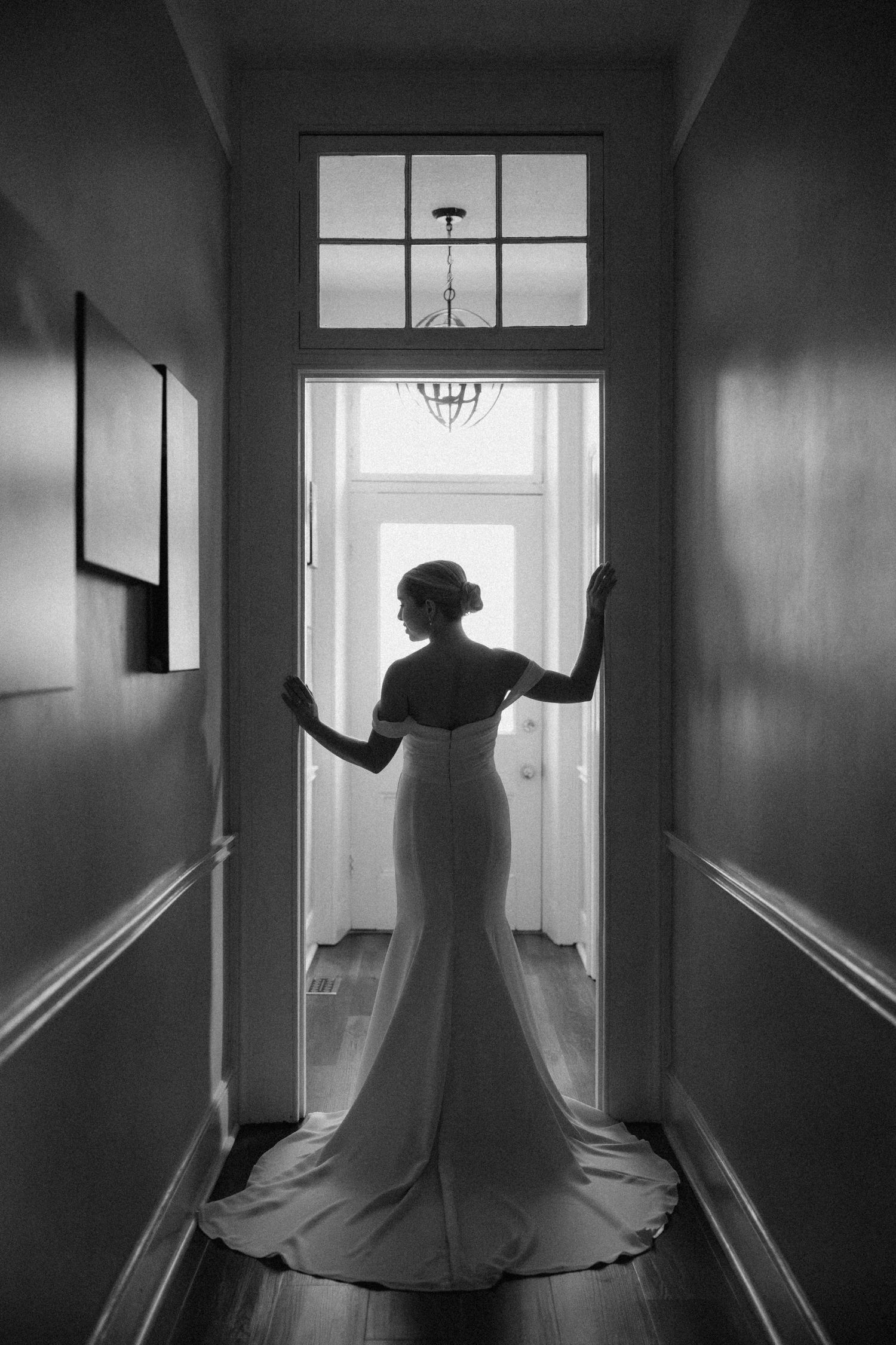 Dramatic black and white silhouette of bride in fitted wedding gown with train standing in hallway doorway.
