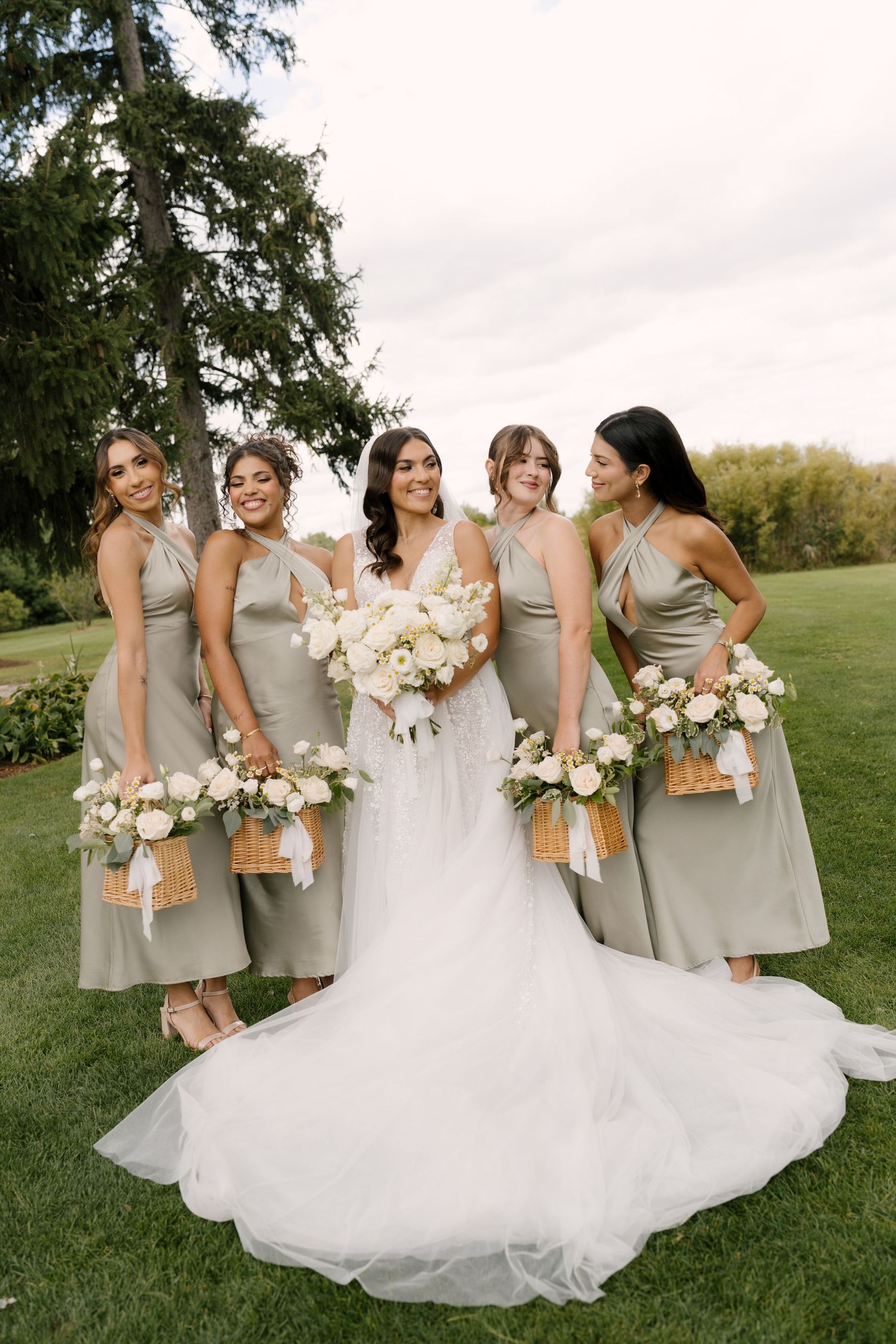 Bride in white lace gown poses with four bridesmaids in sage halter dresses holding wicker basket bouquets.