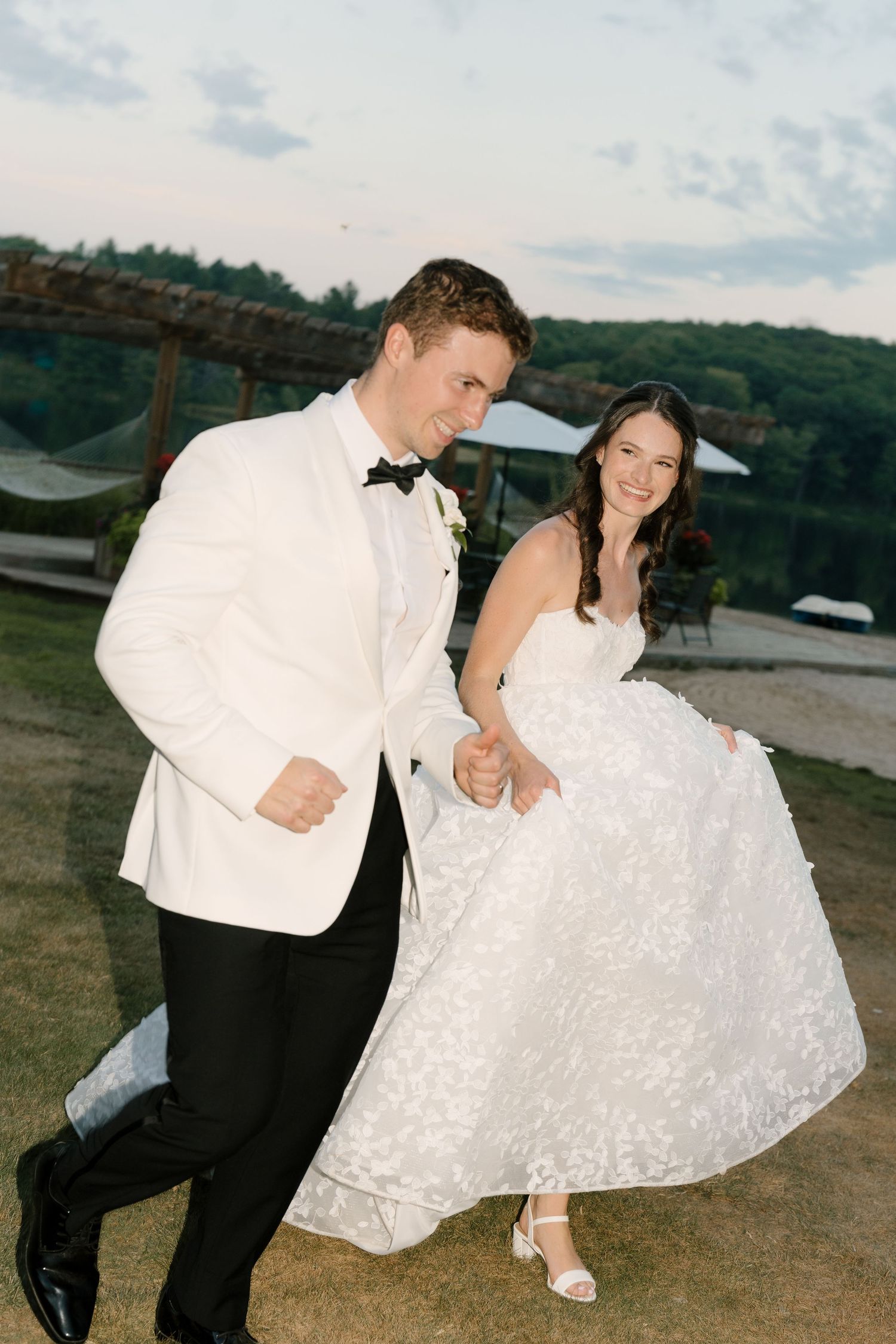 Joyful bride and groom dance outdoors at dusk near a lakeside pergola, bride lifting her floral gown.