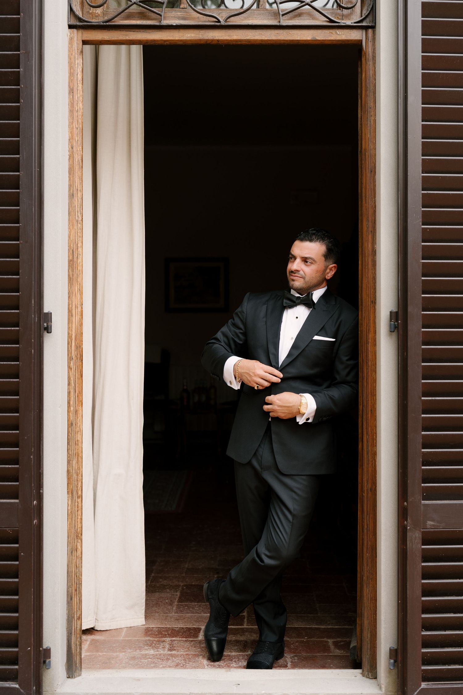 Groom in black tuxedo with bow tie stands in doorway adjusting cufflinks, looking dapper before wedding ceremony.