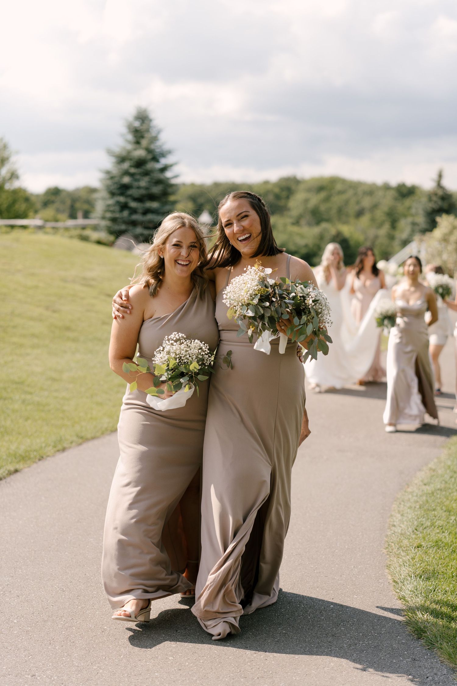 Smiling bridesmaids in taupe gowns walking down a path holding white bouquets at a wedding.