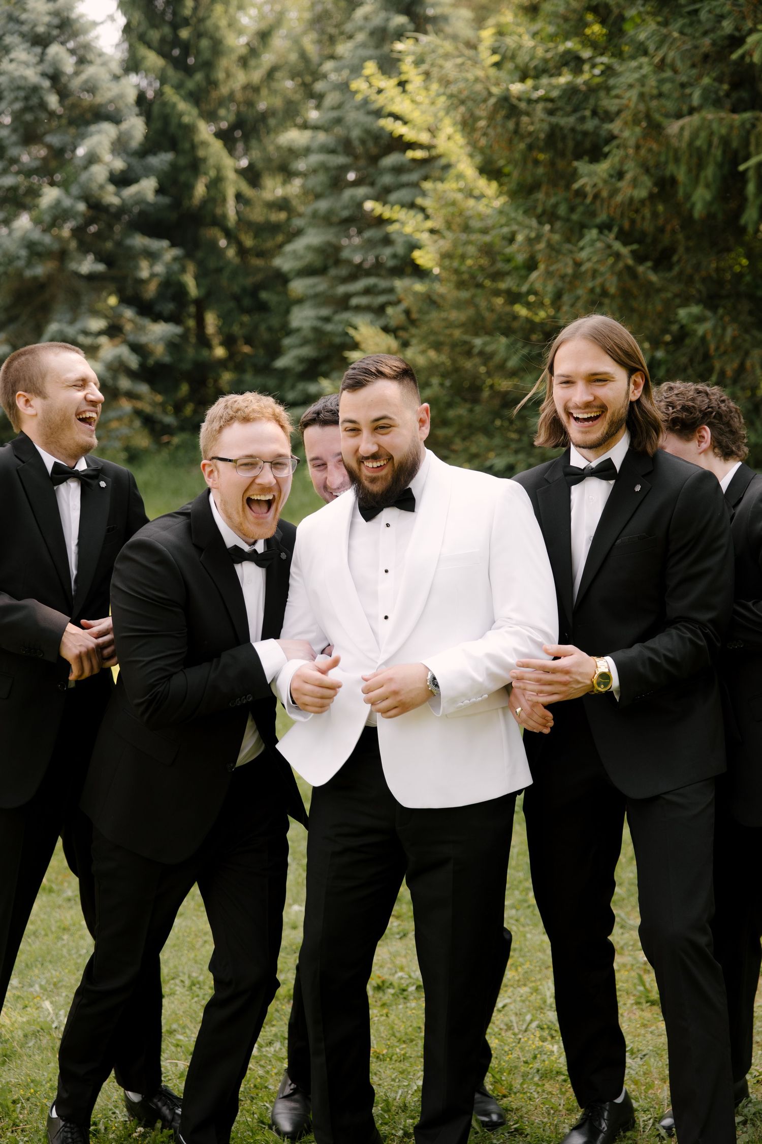 Groom in white tuxedo laughing with groomsmen in black tuxedos outdoors near evergreen trees on wedding day.