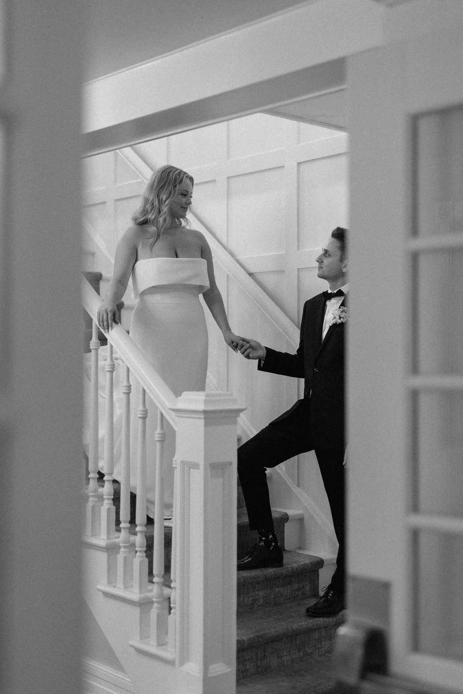 Bride and groom holding hands on Cellar 52 elegant white staircase captured through doorway in black and white.