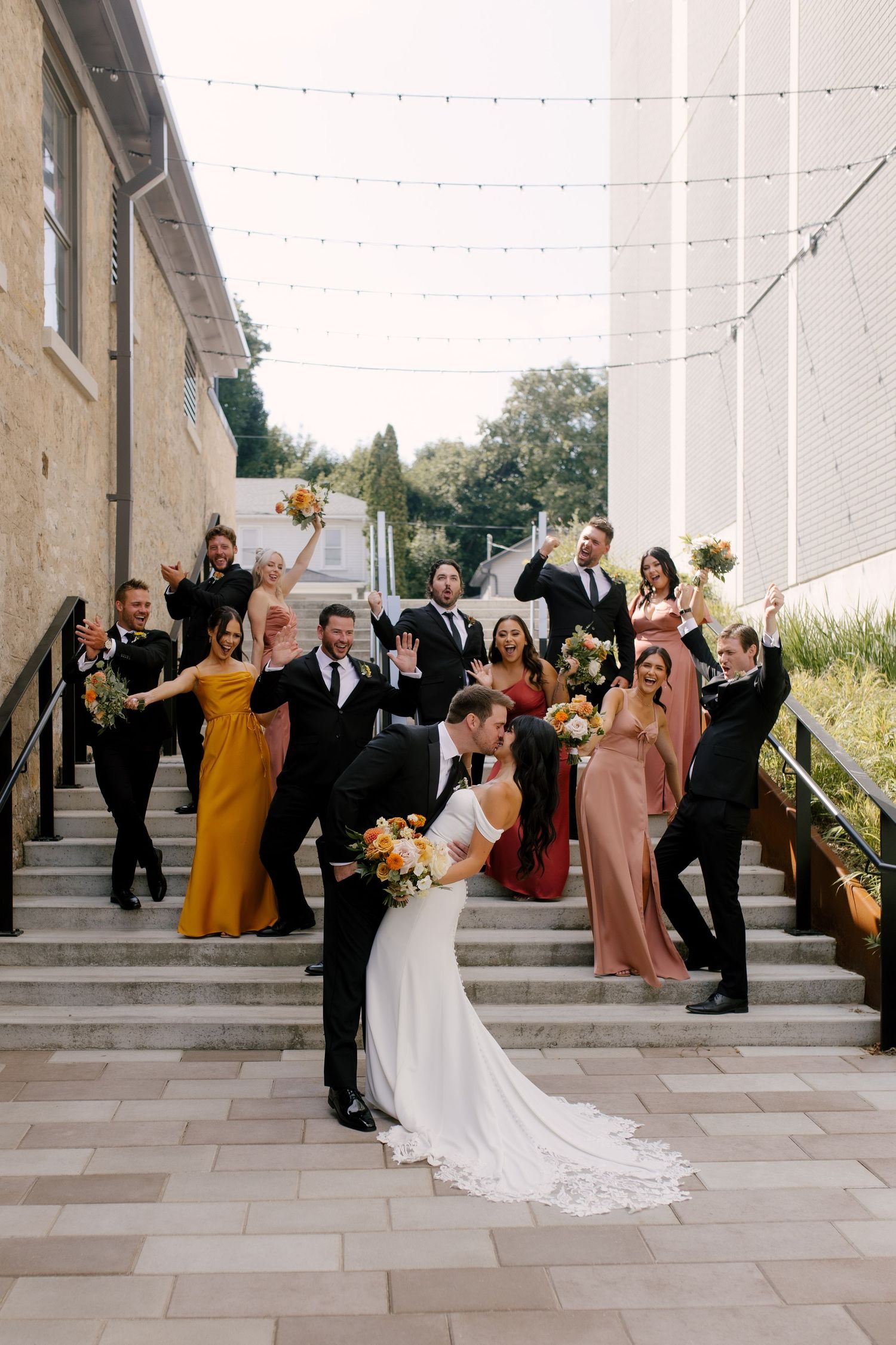 Bride and groom kissing while colourful wedding party celebrates on outdoor stone steps behind them. Located outside Tapestry Hall venue.