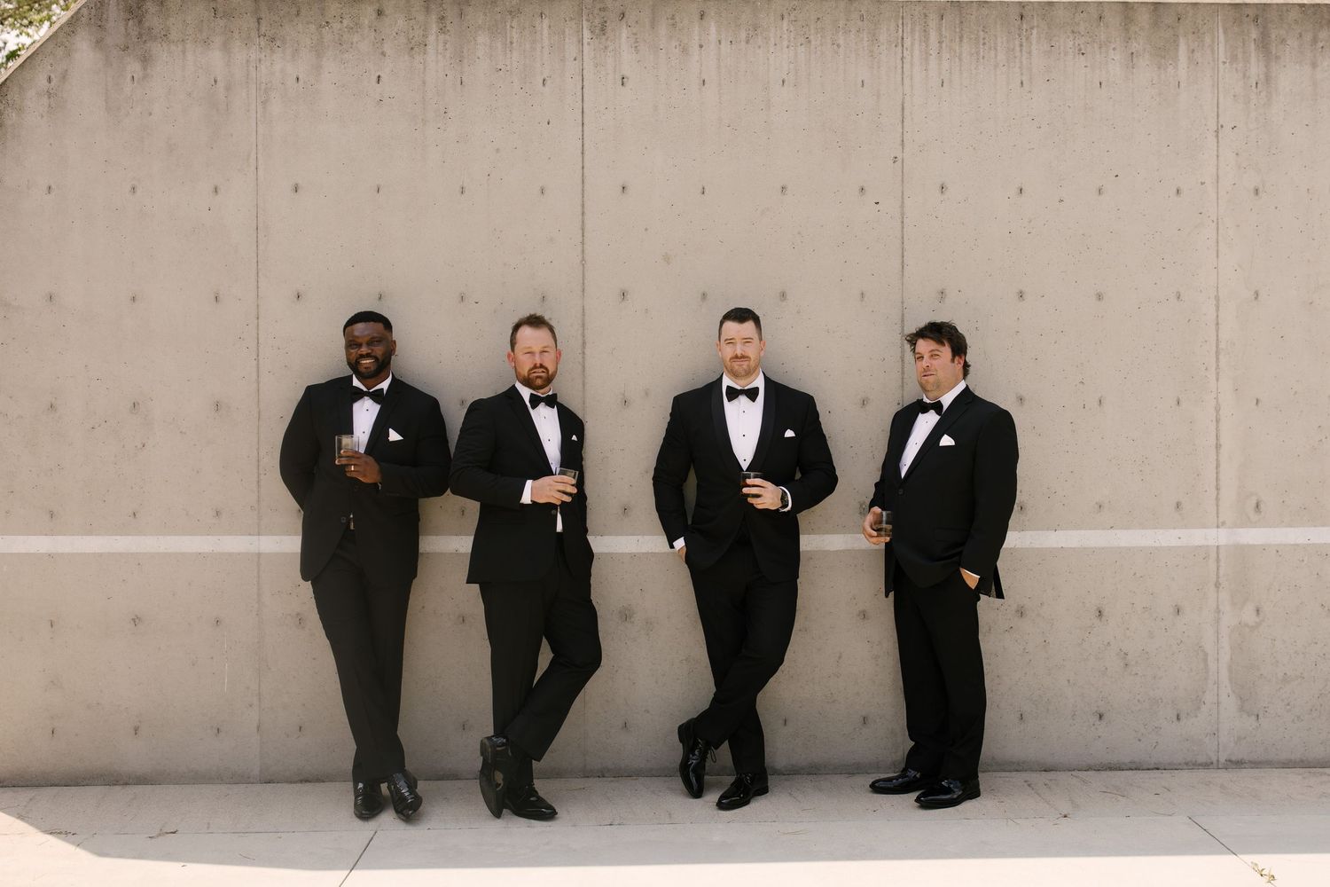 Four groomsmen in black tuxedos with bow ties posing against concrete wall outdoors.