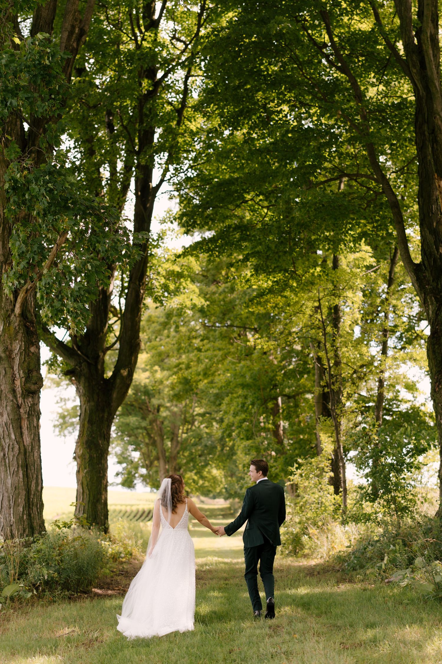 Bride and groom walking hand in hand down a lush green tree-lined path on wedding day.