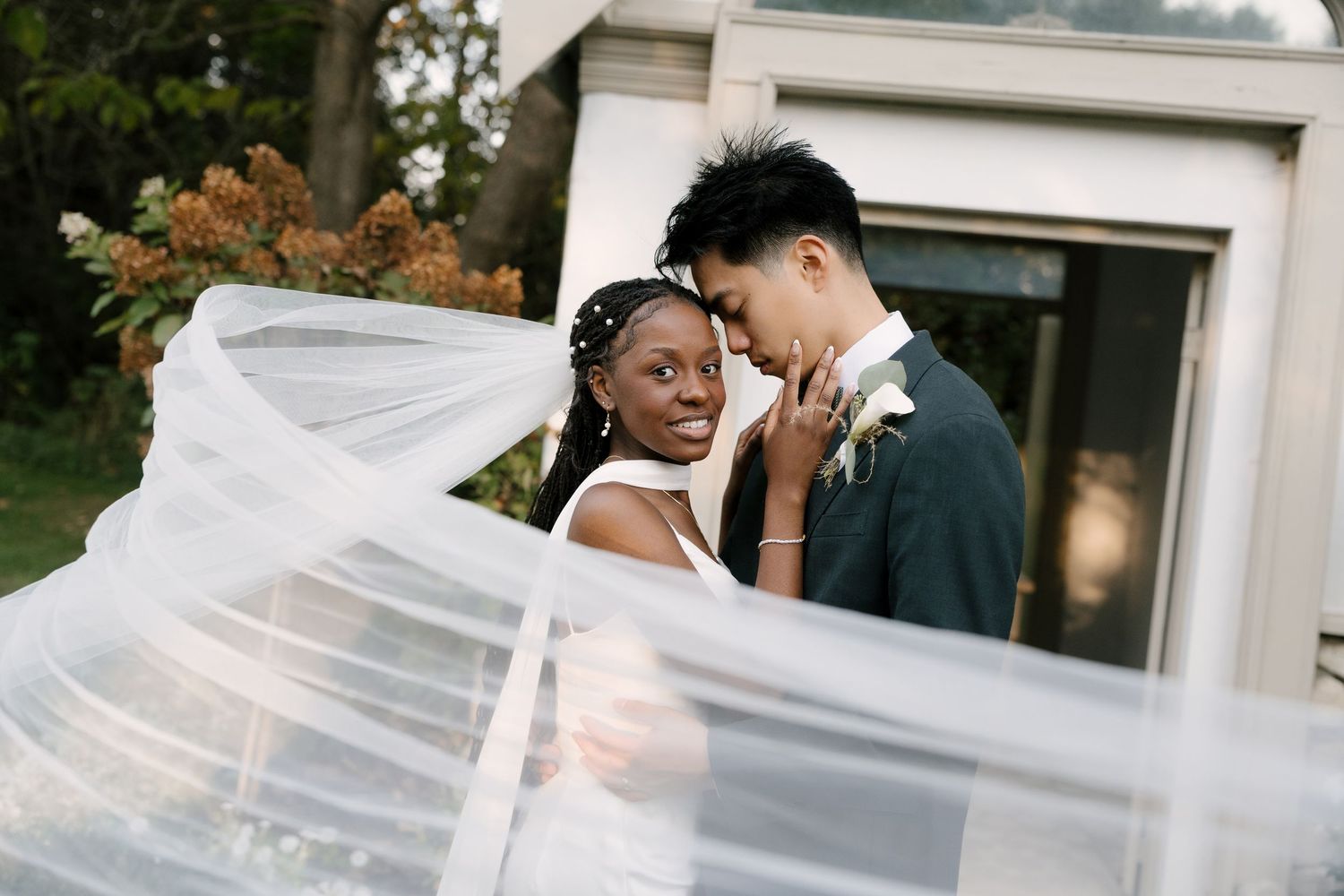 A bride and groom share a romantic moment outdoors, with a flowing veil sweeping across the frame.