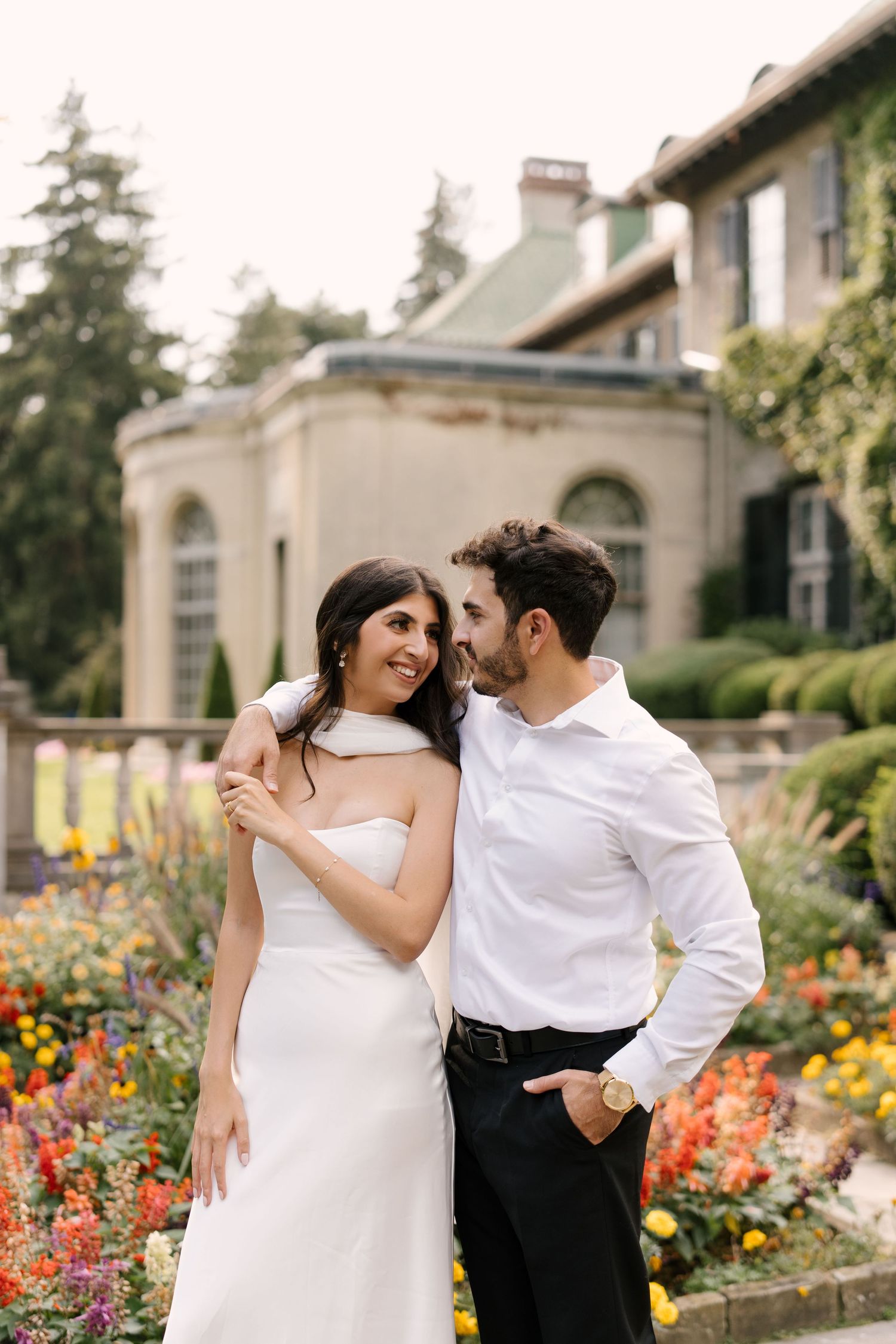 Elegant couple posing in a colorful garden in front of a grand estate, woman in white gown, man in white shirt.