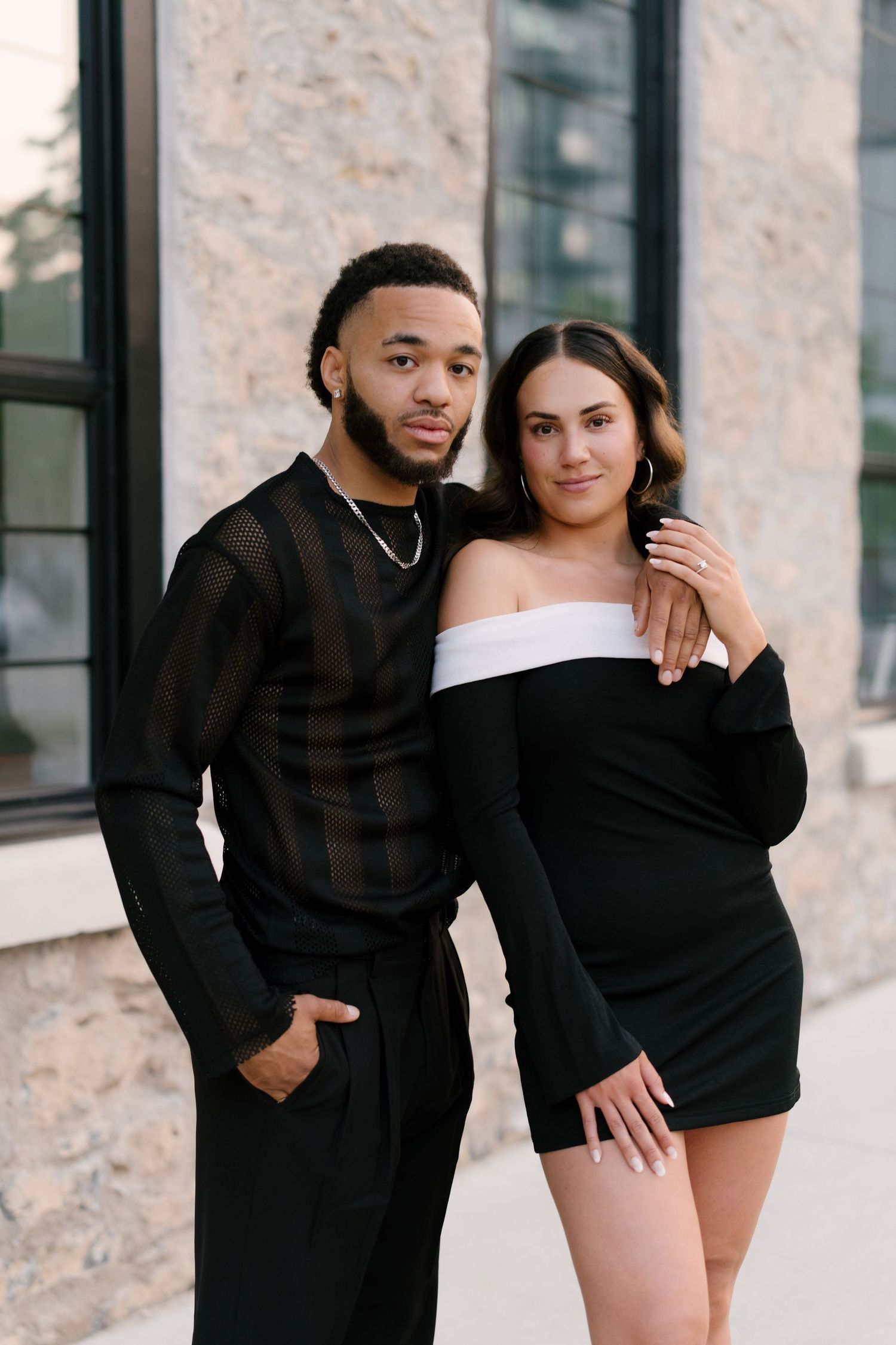 Stylish interracial couple posing together in matching black outfits against a stone building.