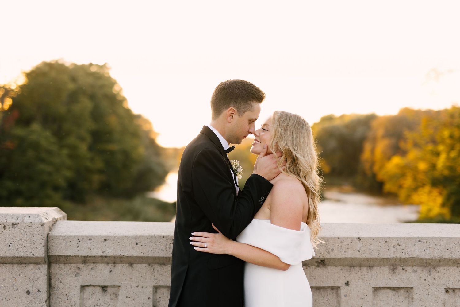 Bride and groom share a tender moment on a stone bridge in St Jacobs surrounded by golden autumn foliage at sunset.