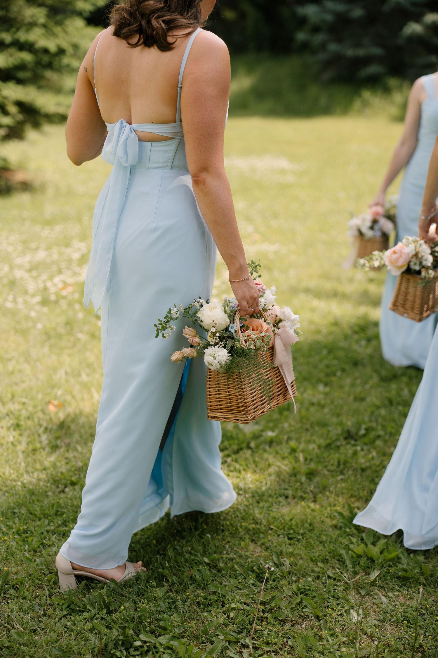 Bridesmaids in light blue gowns walking through green grass carrying wicker baskets filled with pastel flowers.