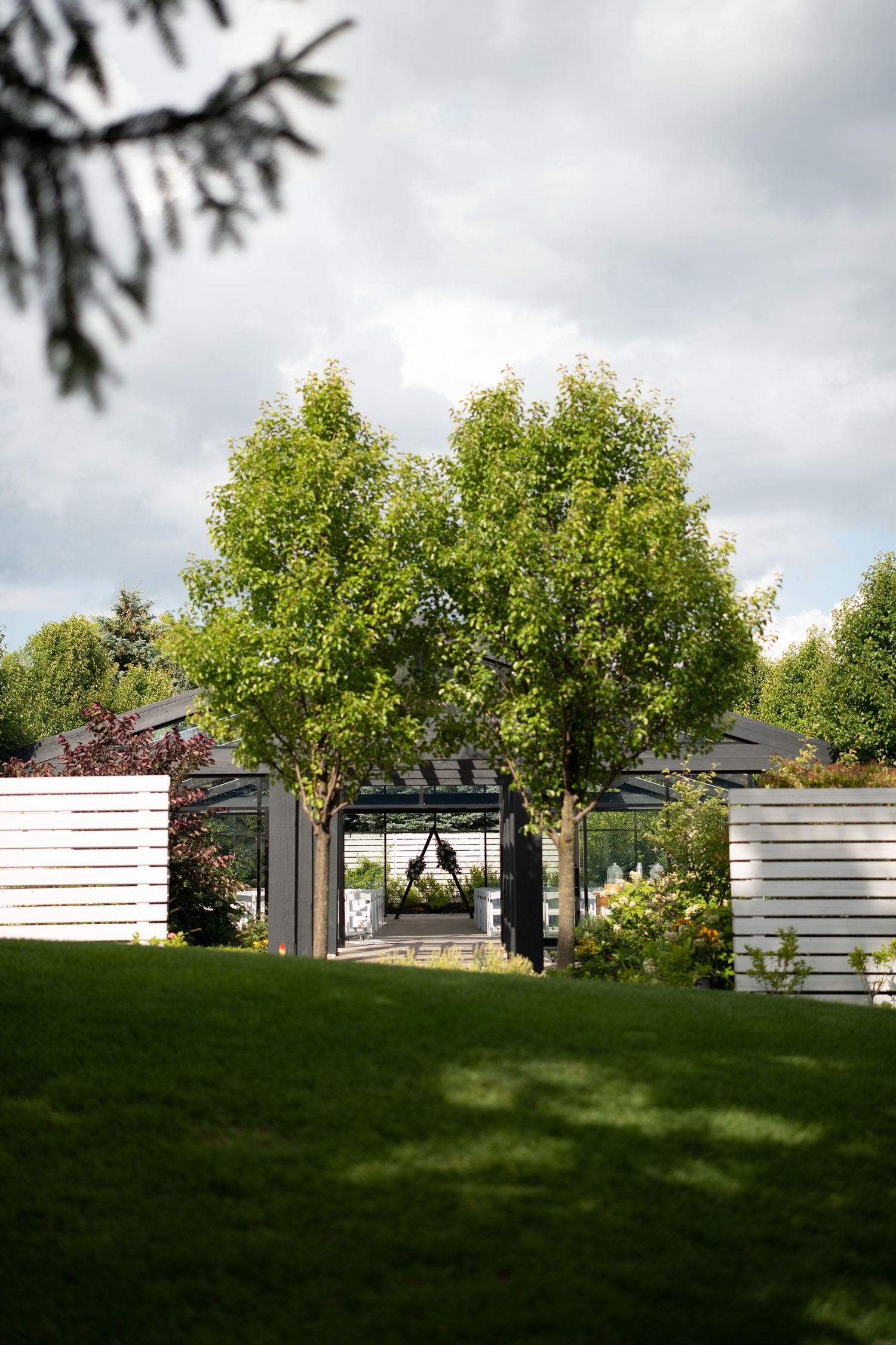 Lush garden Whistle Bear venue with green trees, white fencing, and a black pergola arch under a cloudy sky.
