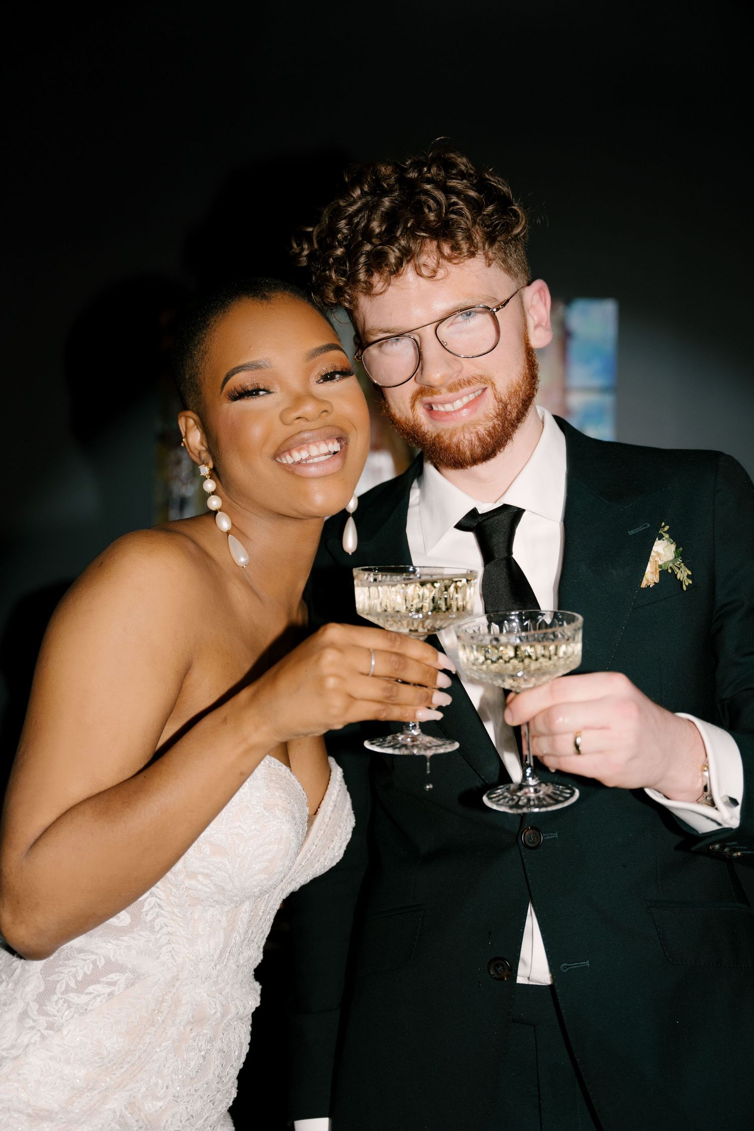 Smiling interracial newlywed couple clinking crystal champagne coupes together at their wedding reception.