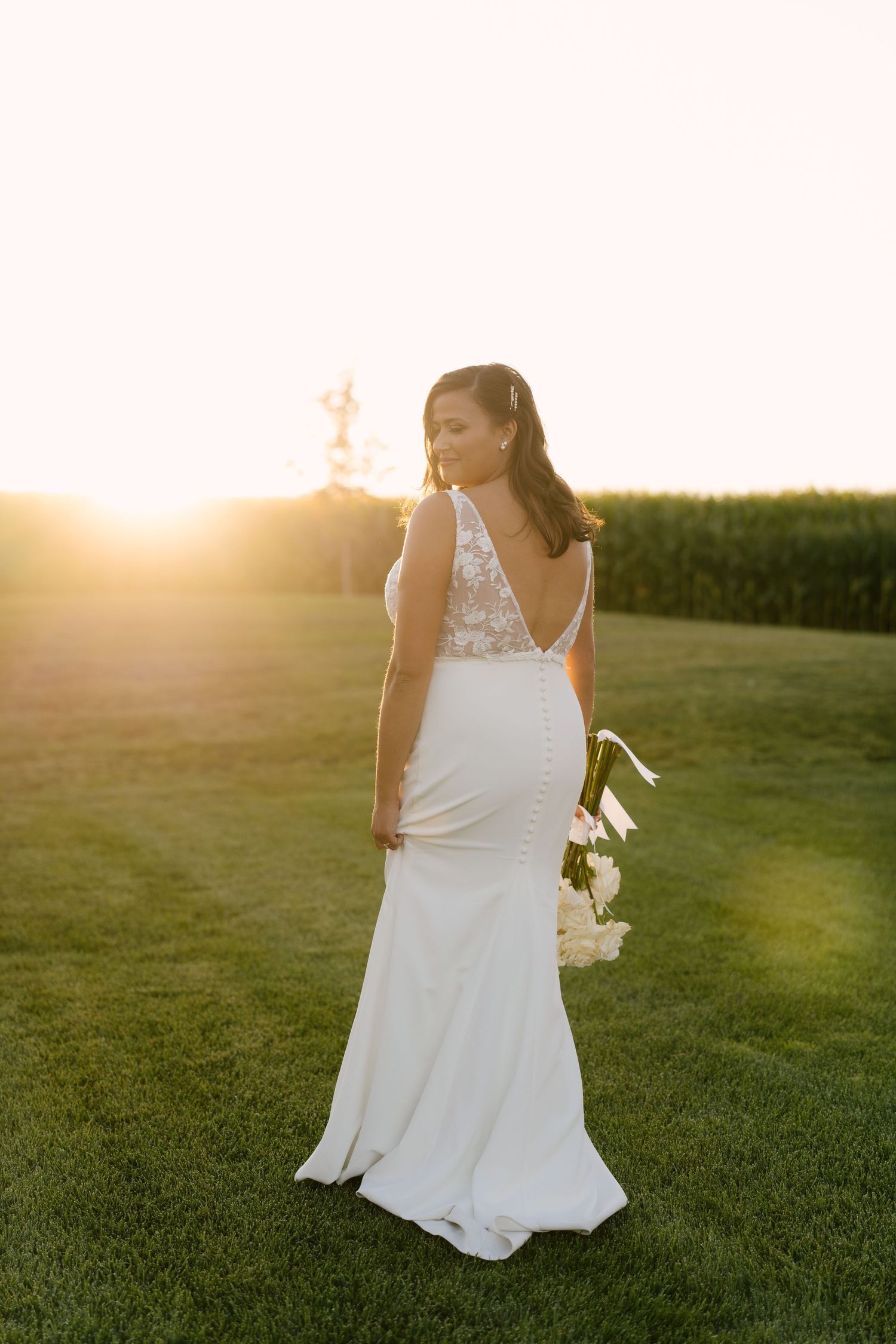 Bride in white lace-back gown holding bouquet stands on green lawn at golden sunset near cornfield.