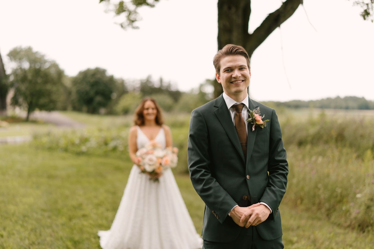 Smiling groom in dark green suit awaits bride during outdoor first look on lush green field at Wakefield Estates.