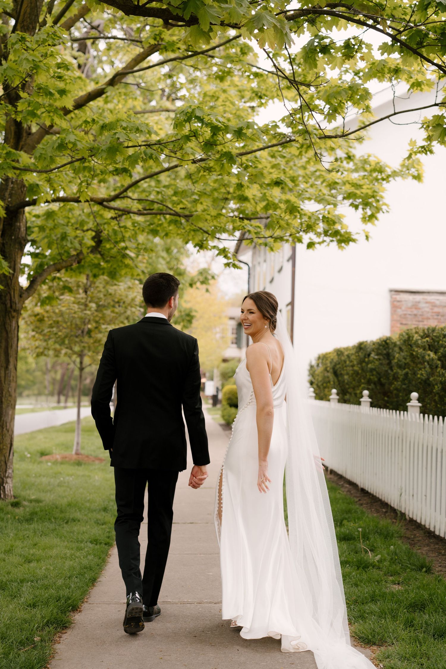 Bride and groom walk hand in hand along a tree-lined sidewalk in Niagara-on-the-lake, bride glancing back in elegant white gown.