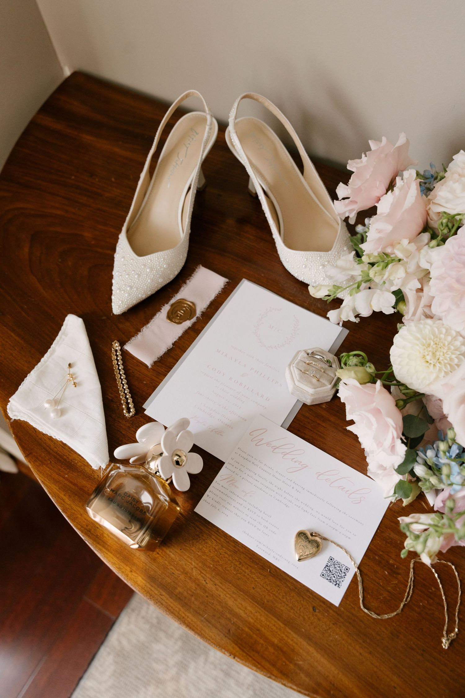 Elegant bridal flat lay featuring white lace heels, floral bouquet, perfume, gold jewelry, and wedding stationery on a wooden table.