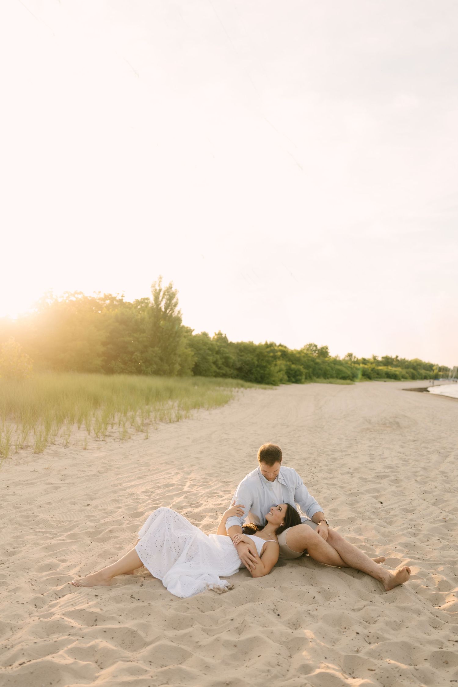 Couple relaxing on sandy beach at golden hour sunset, woman in white dress lying across mans lap.