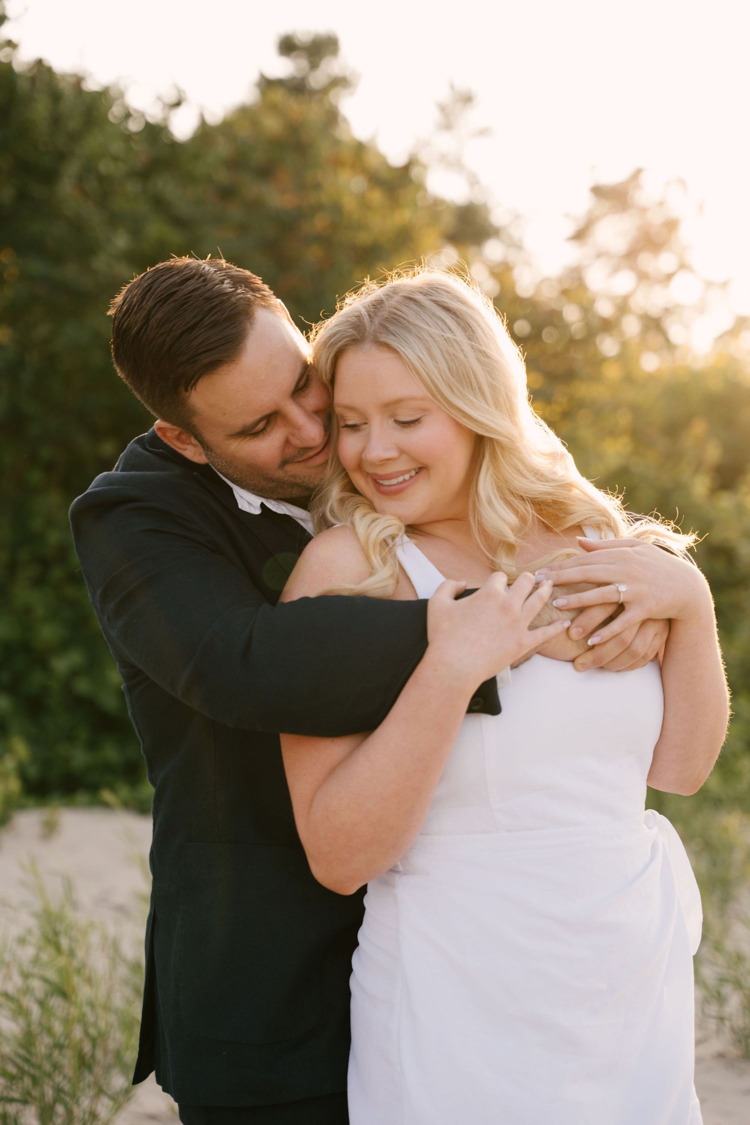Groom in black suit embracing smiling blonde bride in white dress during golden hour outdoor wedding photo.